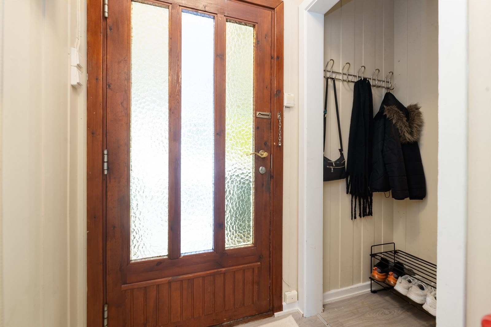 Lite vindfang med lyse overflater og praktisk nisje for oppbevaring av sko- og yttertøy. Eldre ytterdør i tre med glassfelt. Galleribilde