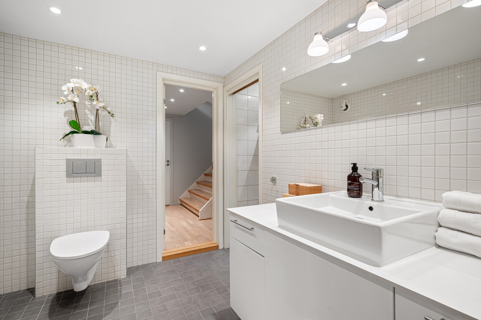 Baderomsinnredning med åpen dusjplass med innfellbare dusjdører, vegghengt wc og servantmontert på benkeplate med underskap og speil. Galleribilde