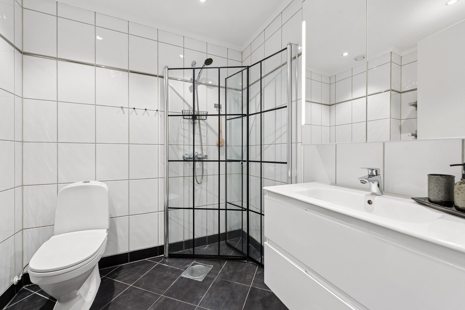 Badet har pene klassiske hvite fliser på vegger og mørke fliser på gulvet. Gulvvarme. Innfellbare dusjdører. Galleribilde