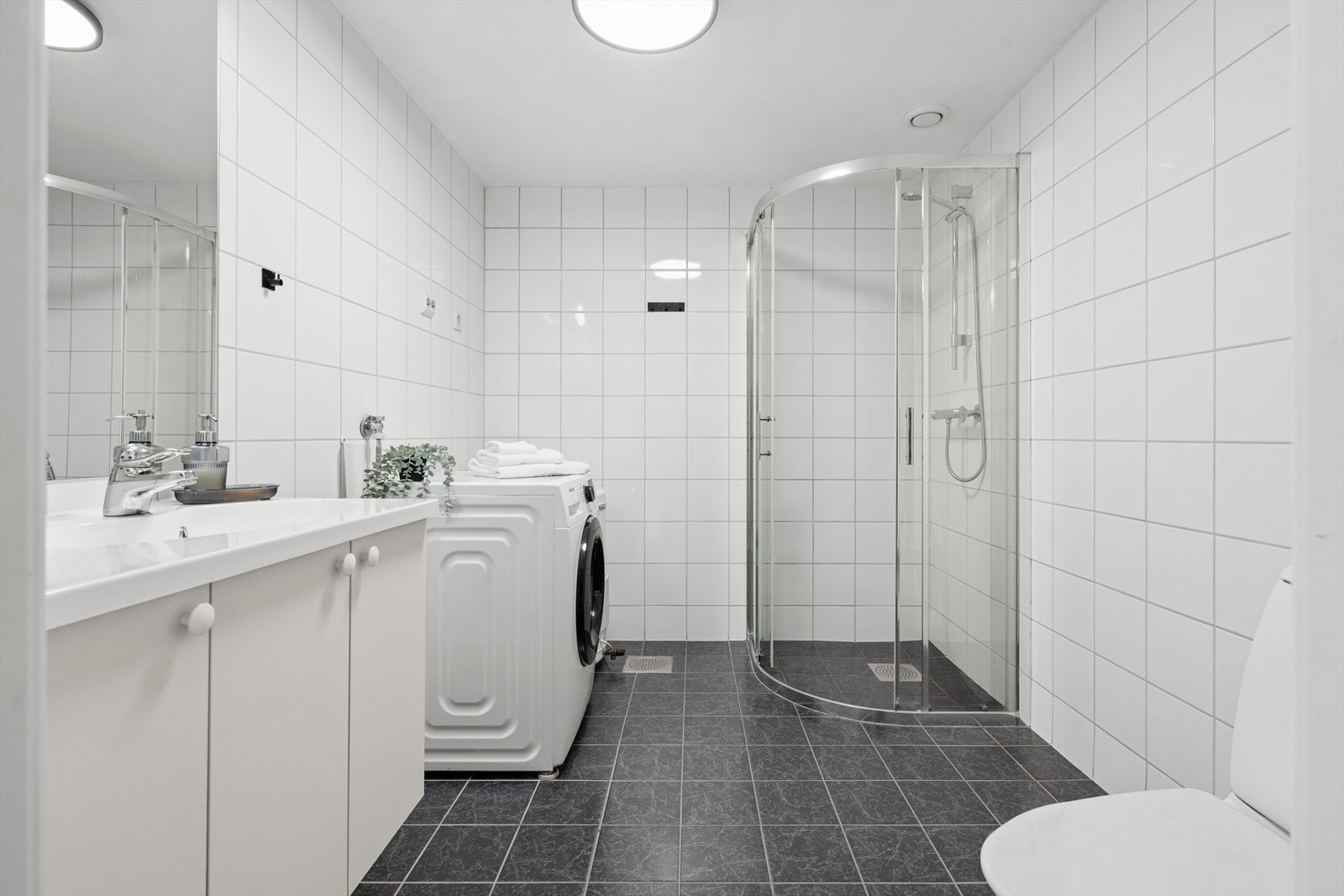 Baderommet er tidløst innredet med grå fliser på gulvet og hvite fliser på vegg. I gulvet har man også varmekabler for ekstra komfort. Badet er ellers utstyrt med dusjhjørne, toalett, baderomsinnredning og opplegg for vaskemaskin. Galleribilde