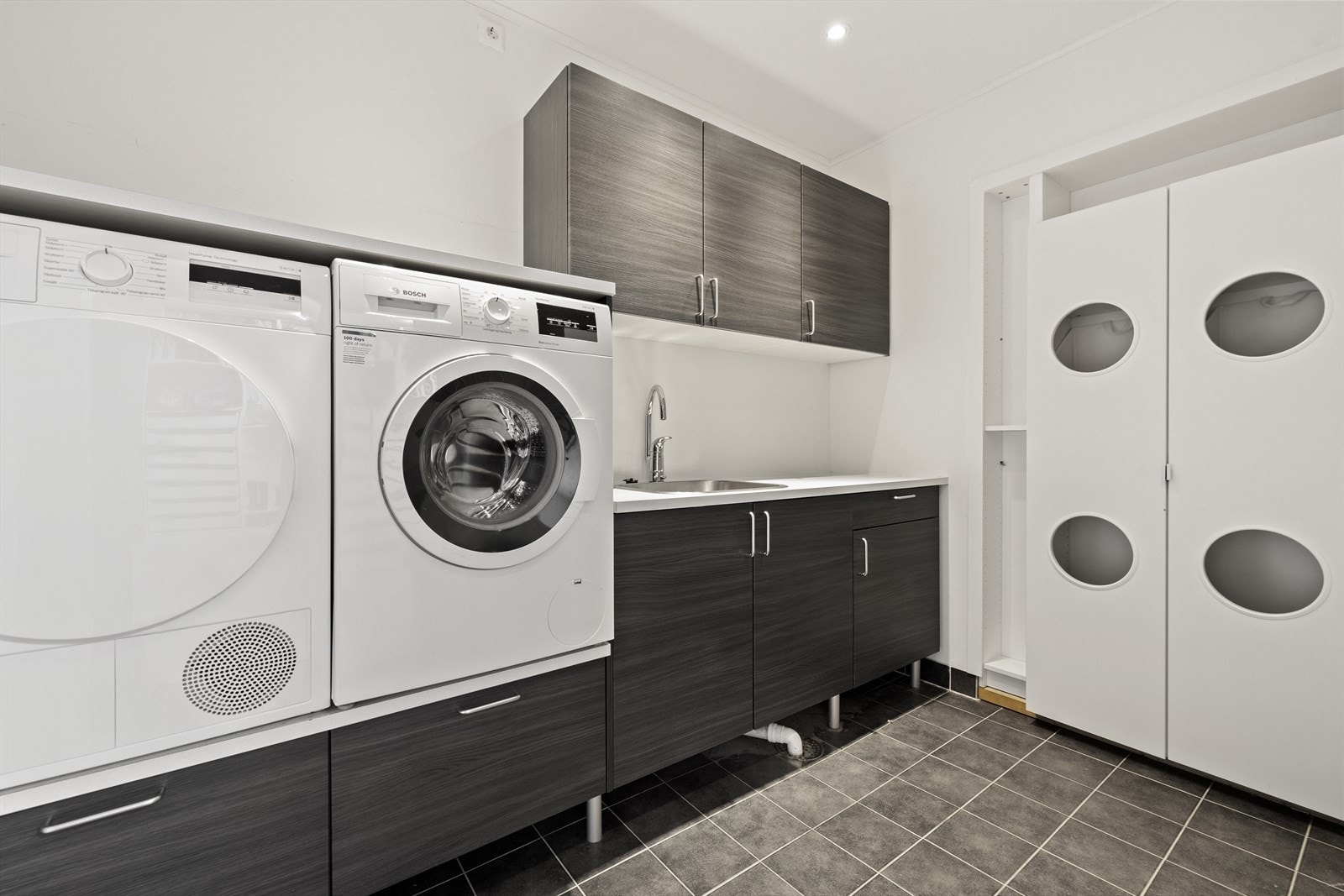 Praktisk vaskerom med flislagt gulv, god innredning og rikelig med oppbevaringsplass. Tilrettelagt for både vaskemaskin og tørketrommel. Galleribilde