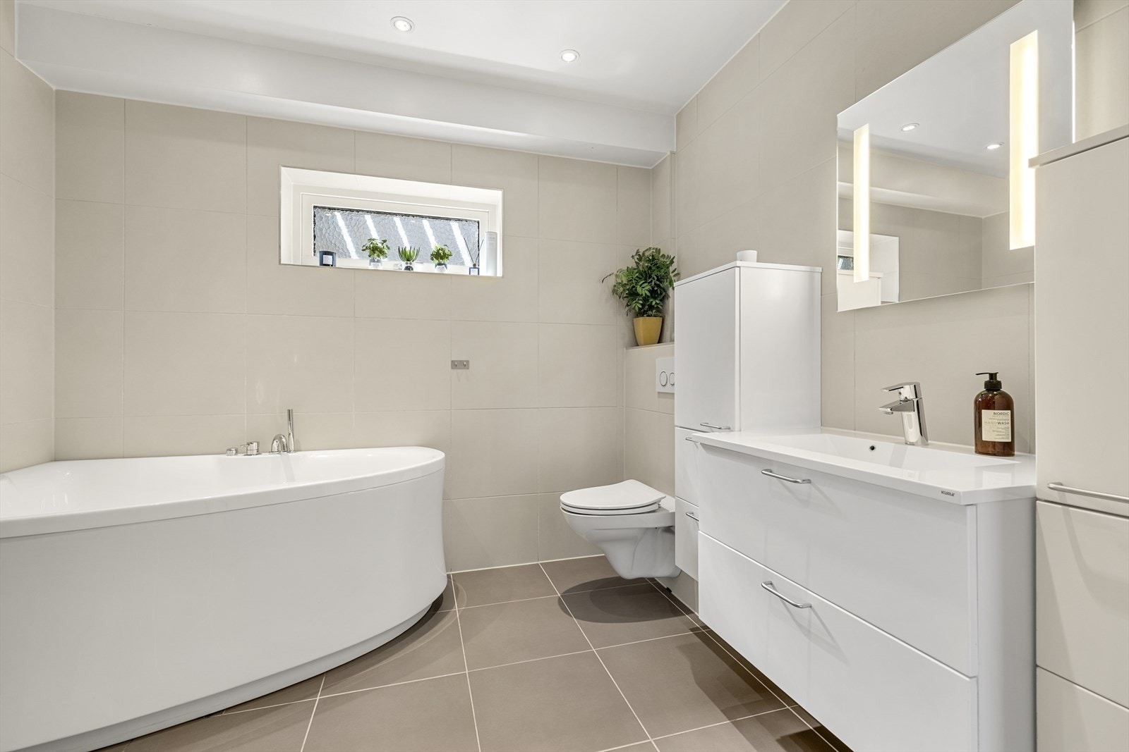 Pent, flislagt baderom i underetasjen, holdt i tidløse fargetoner og utstyrt med vegghengt toalett, servant med skuffer og badekar. Galleribilde