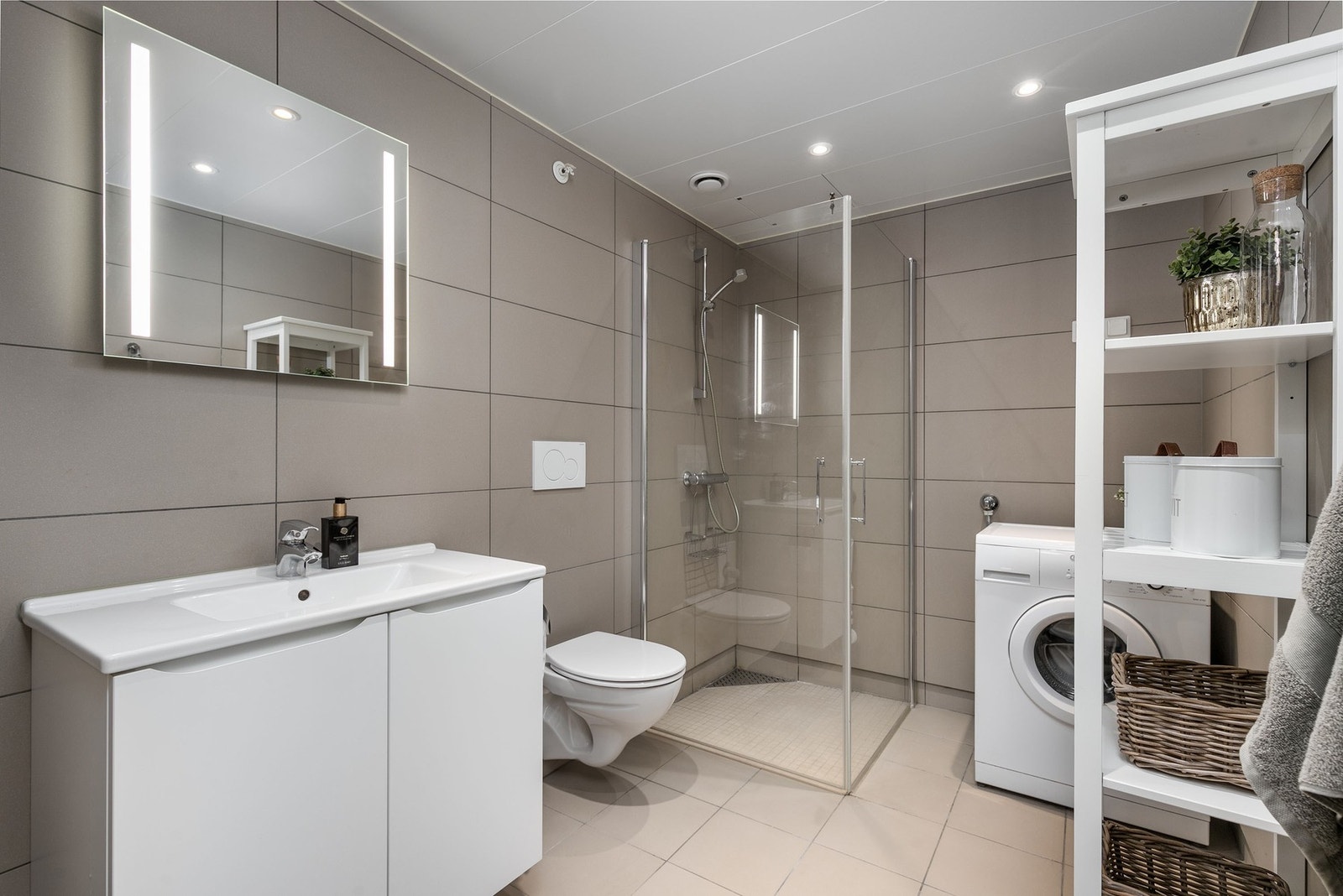 Badet er fra byggeår og fremstår lyst og moderne, med flislagte vegger og malt himling med downlights. Innredningen består av servant med glatte fronter og speil, vegghengt toalett og dusjsone med dusjvegger. Det er opplegg for vaskemaskin. Galleribilde