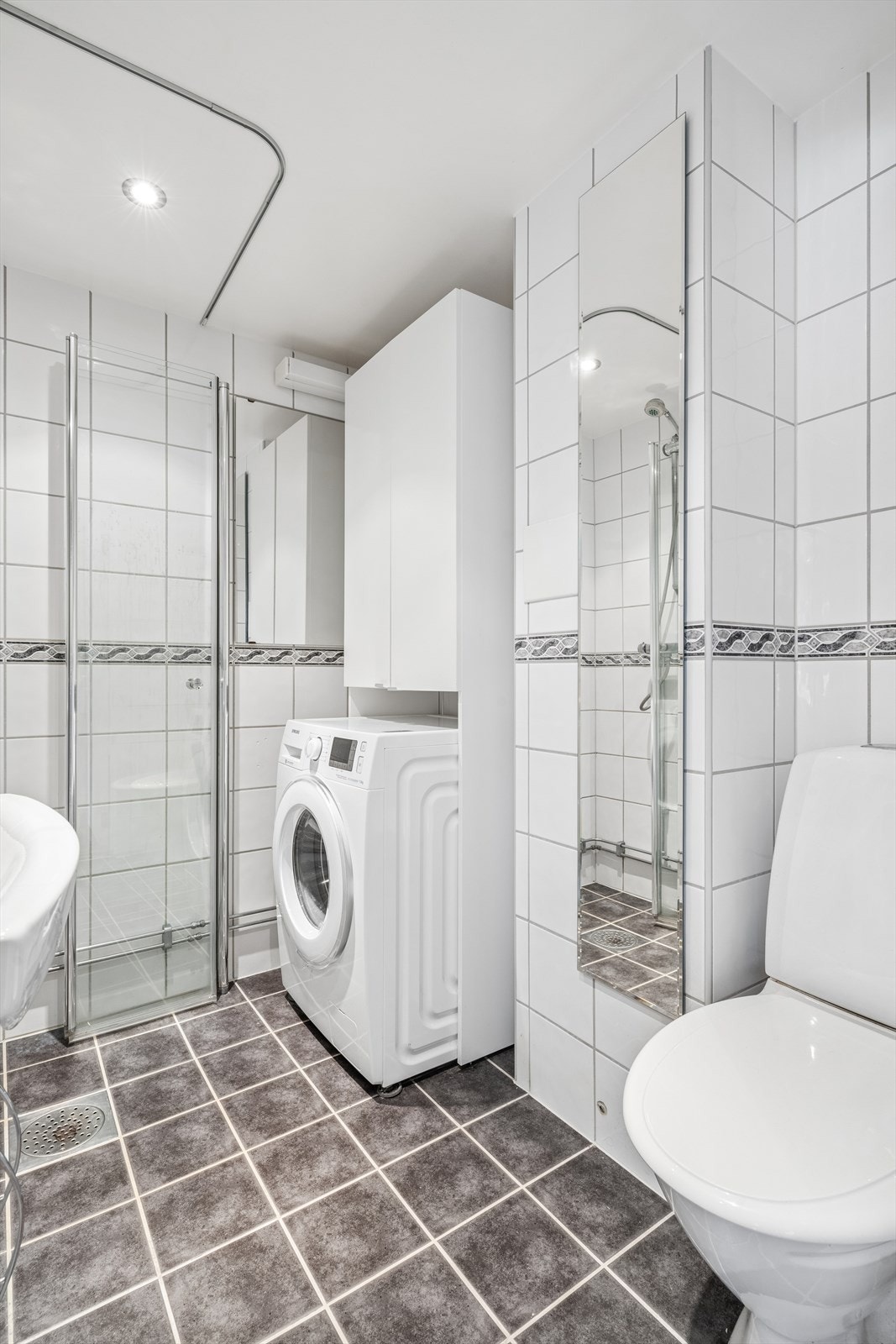 Baderomsinnredningen består av gulvmontert toalett, servant med underskap, speilskap, og dusjnisje med innfellbare glassdører Galleribilde