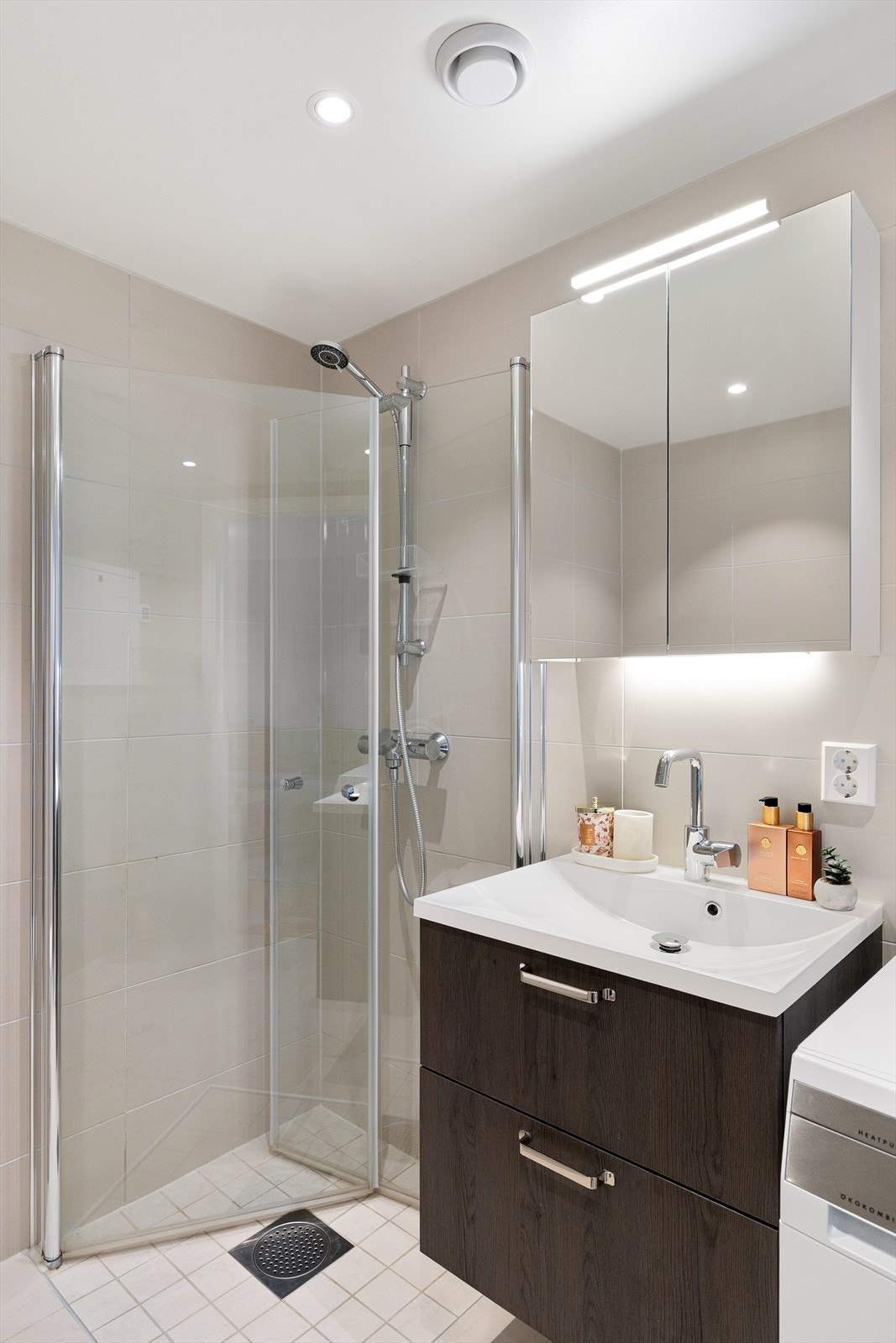 Badet er utstyrt med veggmontert toalett og dusjhjørne med innfellbare dører i herdet glass. Baderomsinnredning med skuffer, ovenpåliggende servant med ett-greps armatur, vegghengt speilskap og belysning. Det er opplegg for vaskemaskin. Galleribilde