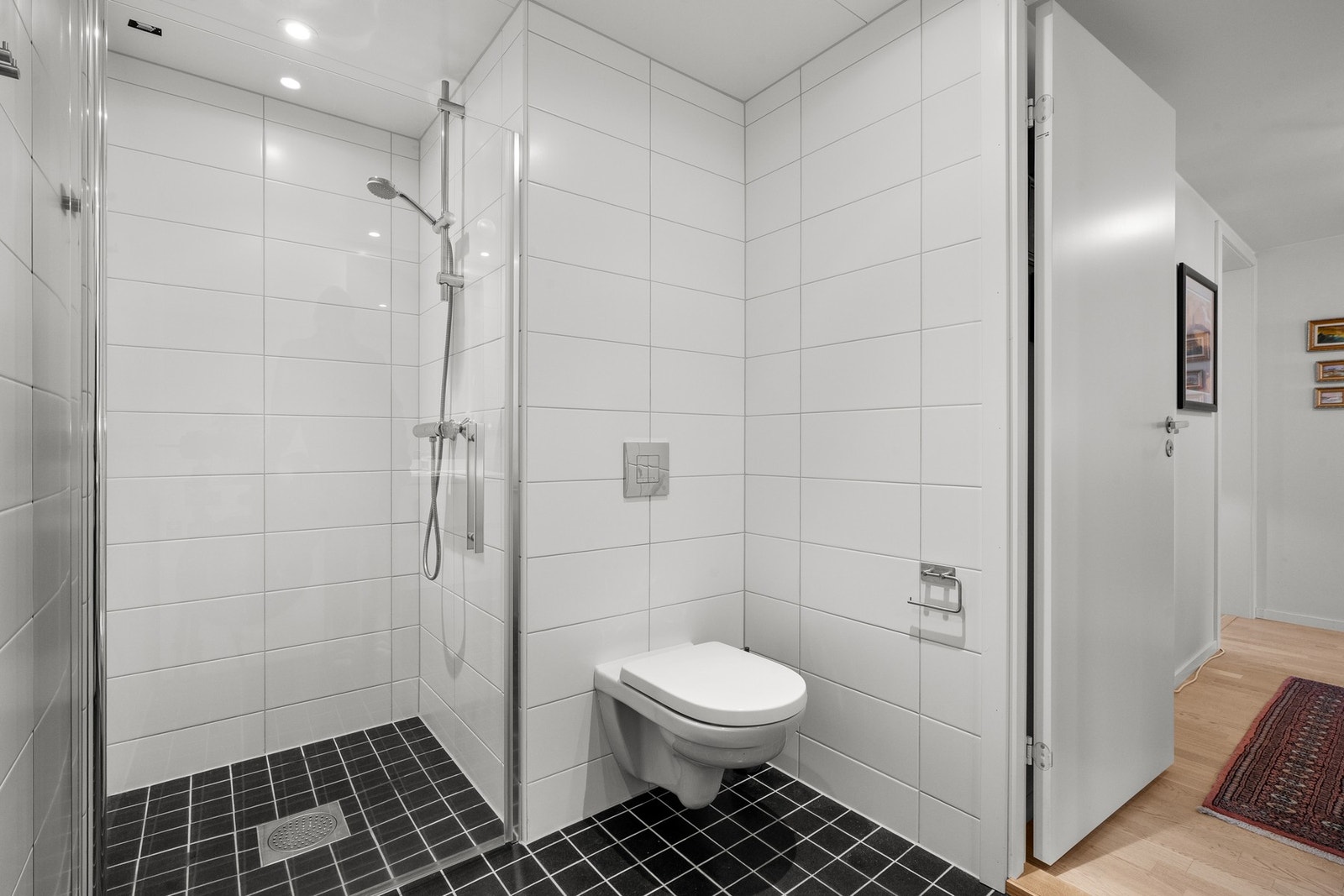 Baderommet er også utstyrt med dusjnisje med glassdør og vegghengt toalett. Baderommet har mørke fliser på gulv, elektrisk gulvvarme, hvite fliser på veggene, downlights i himlingen og avtrekk via balansert ventilasjon. Galleribilde