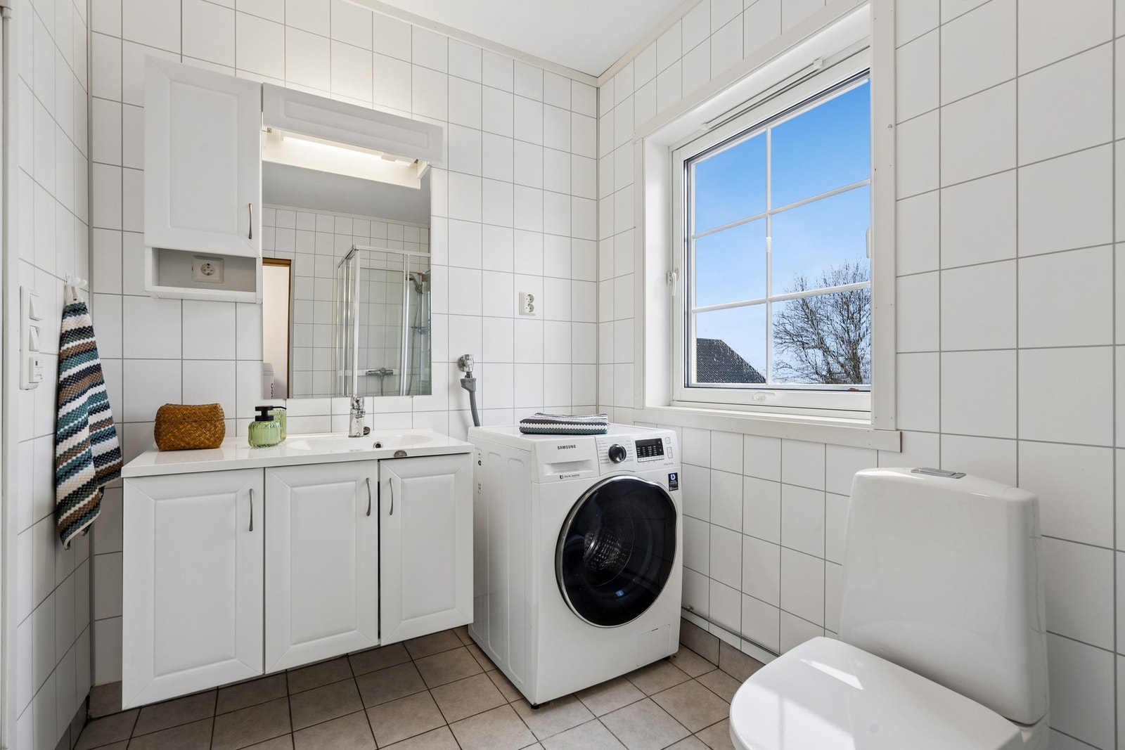 Bad i første etasje er flislagt med opplegg for vaskemaskin. Rommet har et stort vindu som slipper inn mye naturlig lys Galleribilde