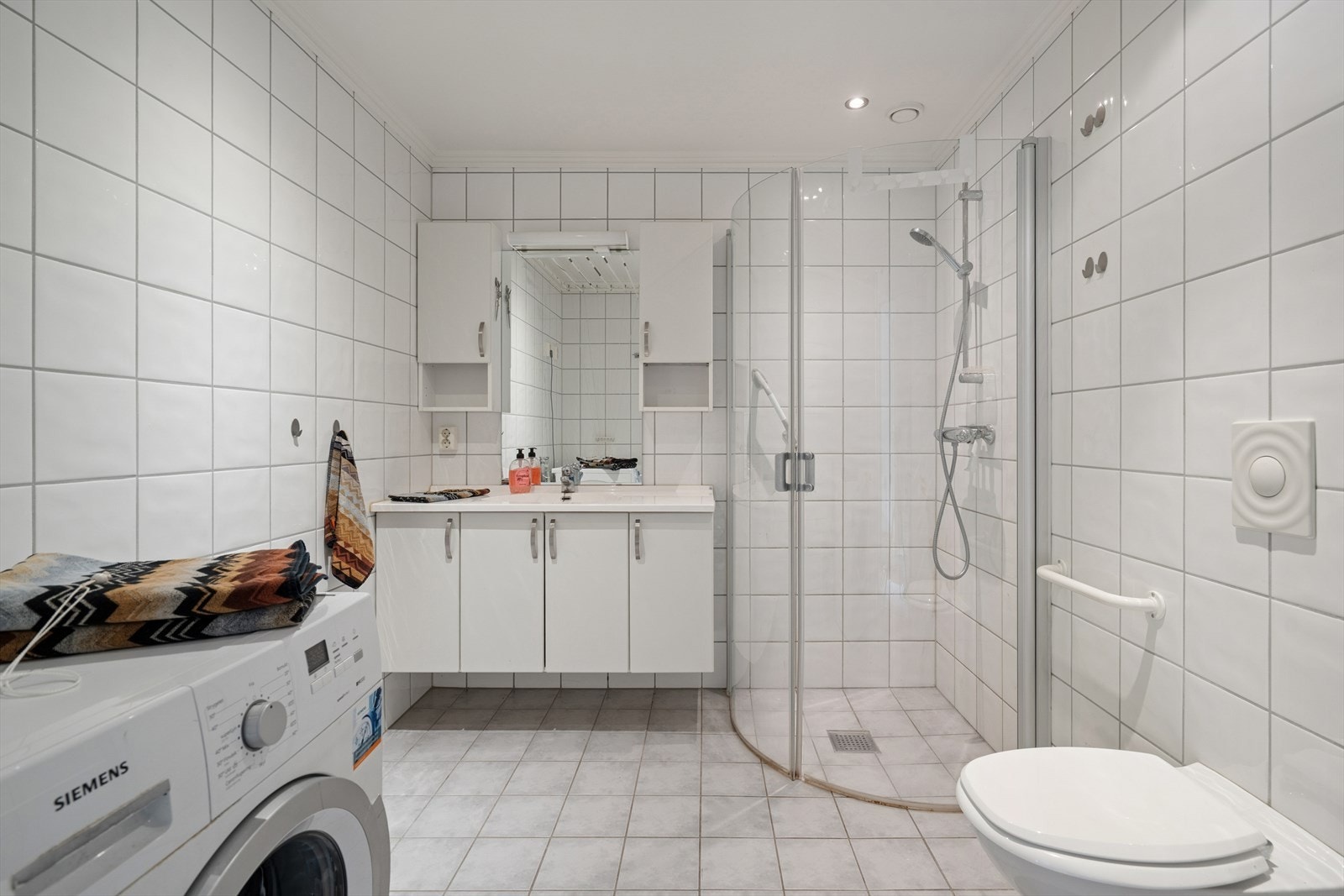 Badet er flislagt med varmekabler i gulv og inneholder baderomsinnredning med nedfelt servant, vegghengt speil, vegghengt toalett og dusjhjørne. Opplegg for vaskemaskin. Galleribilde