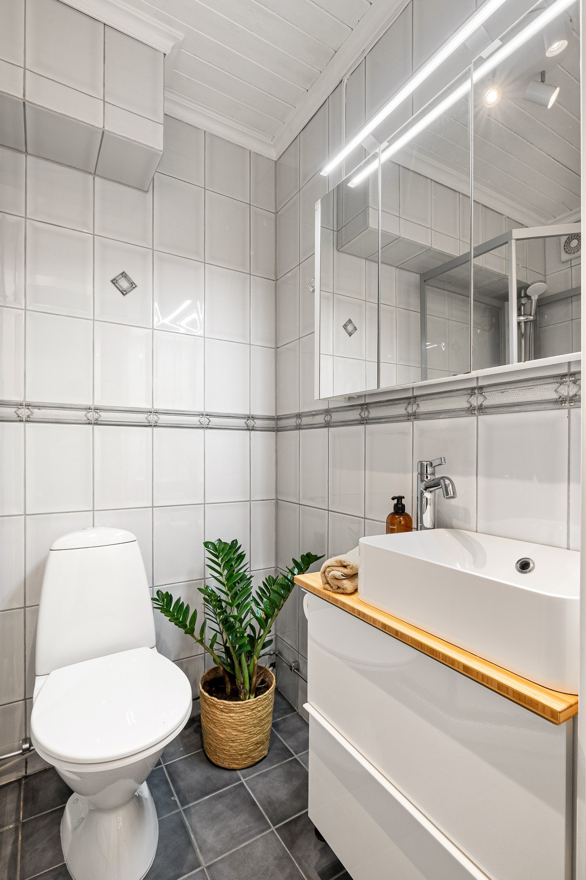 Badet inneholder servant på underskap, speilskap med belysning, kran og avløp for vaskemaskin, dusjkabinett og gulvmontert WC. Galleribilde