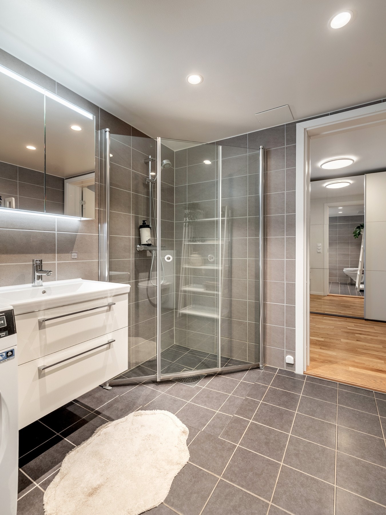 Innredningen består av servantinnredning, speil i dører på overskap på vegg over servant, vegghengt toalett, dusjhjørne med svingbare glassdører og opplegg for vaskemaskin. Galleribilde