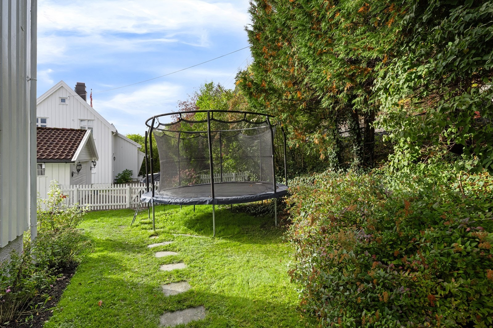 Tomten er frodig og grønn på sommeren med trampolineplass Inngjerdet hage med port og busker. Galleribilde