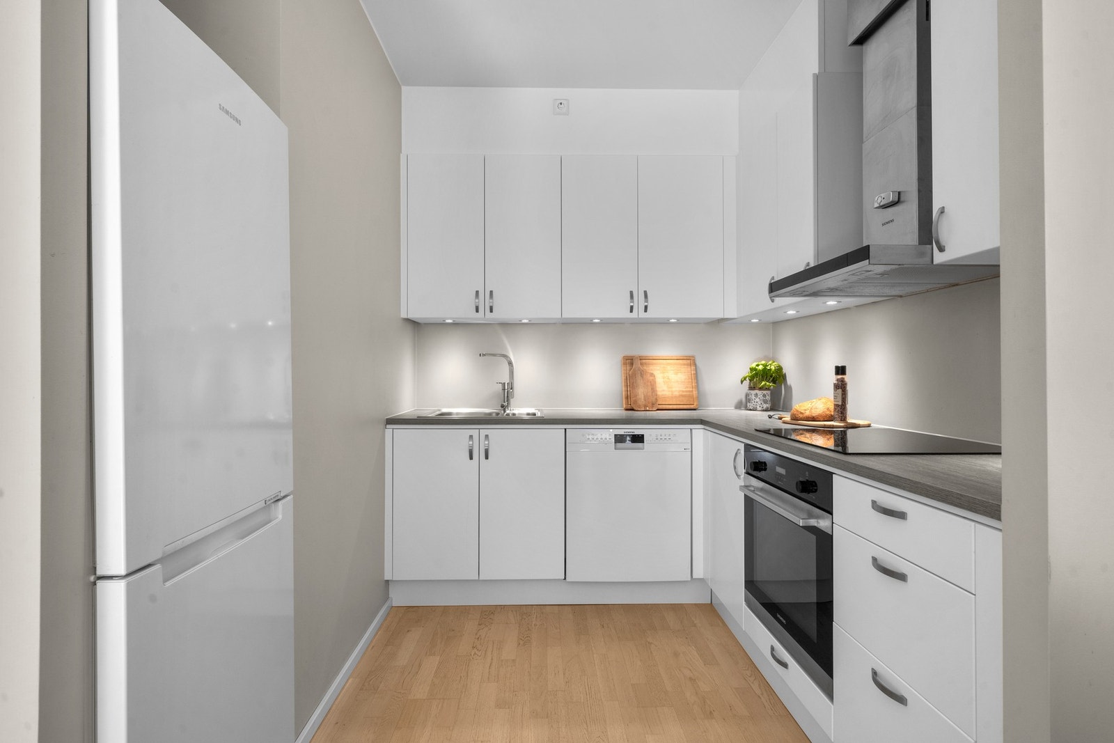 Kjøkkeninnredningen er plassert i en L-form, pluss en nisje til kjøleskap langs den tredje veggen. Innredningen har takoppforede skap, slette fronter med smale grep, samt benkeplater i laminat med nedfelt vaskekum i stål. Galleribilde