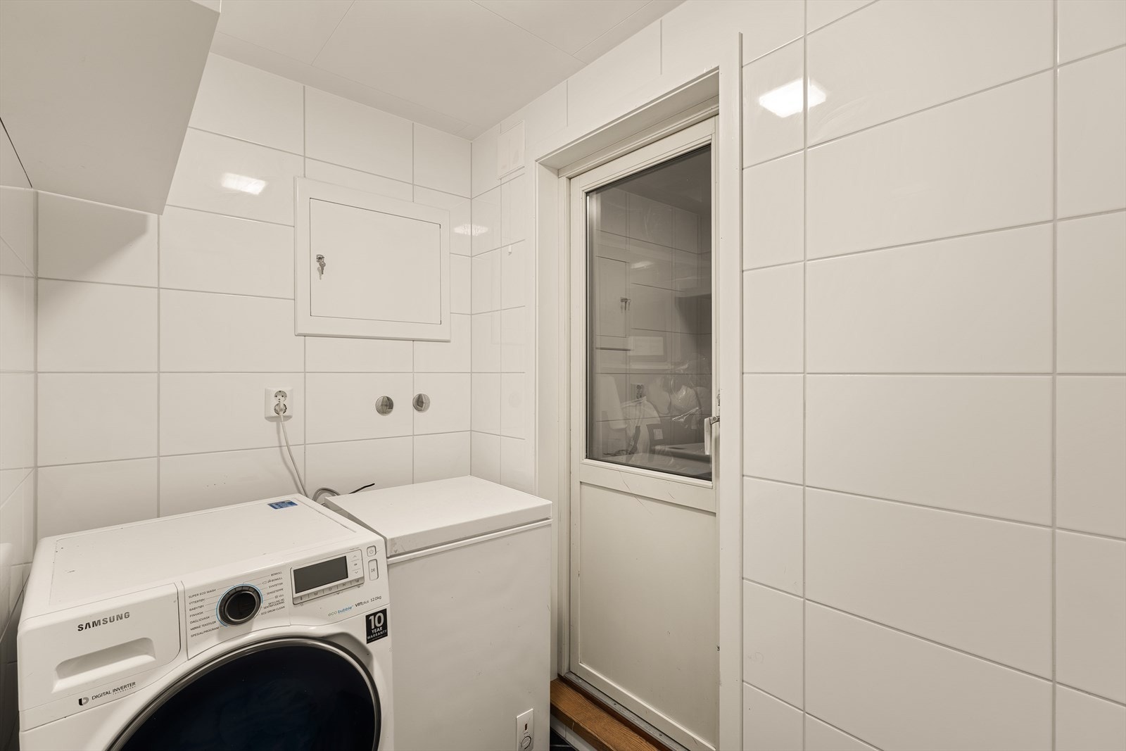 Et romslig vaskerom som er praktisk innrettet med opplegg for vaskemaskin. Rommet har keramiske fliser på gulv og vegger og takplater i himling. Her er det også plass til ekstra fryseboks ved behov Galleribilde