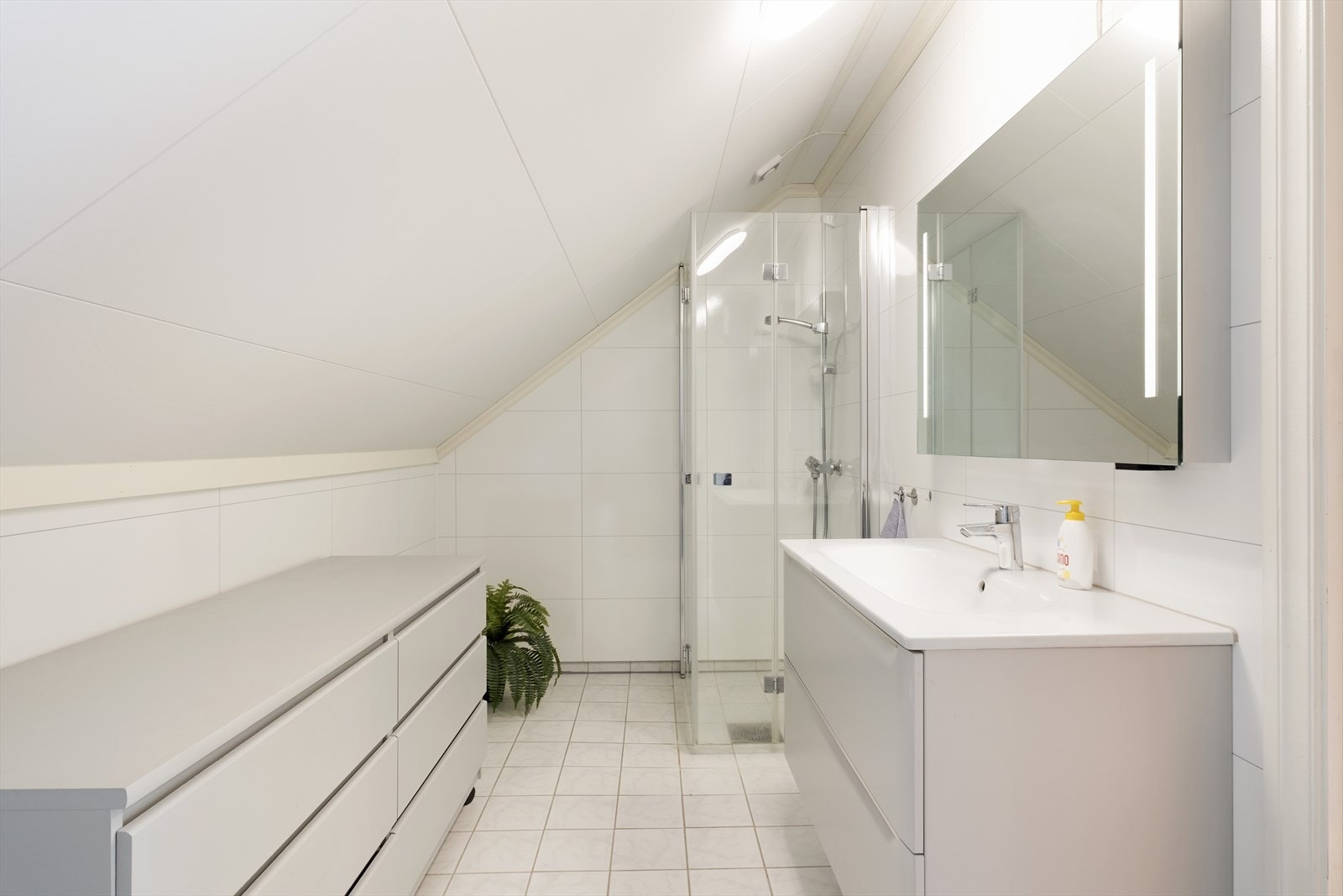 Badet har flis på golv og baderomsplater på veggar. Rommet inneheld golvmontert toalett, servantskap med heilstøypt servant , speigel med integrert belysning og dusjnisje med foldedørar i glas. Galleribilde