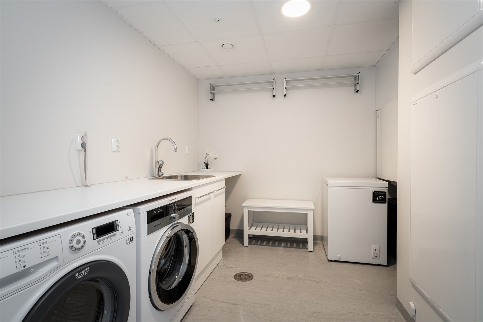 Leiligheten har et funksjonelt kombinert vaskerom og teknisk rom med belegg og sluk i gulv. Hvitmalte vegger og moderne letthimling med aluminiumsprofiler skaper et ryddig uttrykk. Rommet er innredet med underskap, benkeplate og nedfelt utslagsvask. Galleribilde