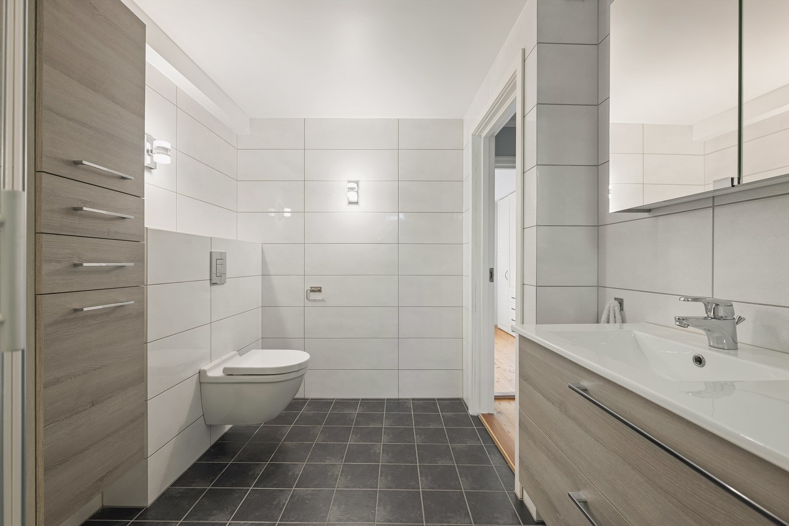 Innredningen består av servant på benkeskap, speilskap med belysning og et dusjkabinett med skyvedører. Toalettet er veggmontert med innbygd sisterne, noe som gir et ryddig uttrykk og enklere renhold. Galleribilde