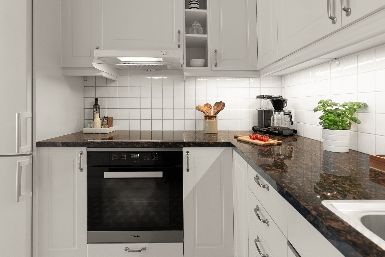 Kjøkkeninnredningen er utstyrt med hvite, profilerte fronter, benkeplate i steinkompositt med planlimt kum i rustfritt stål og flisfelt på vegg over benk, samt belysning under overskap. Galleribilde