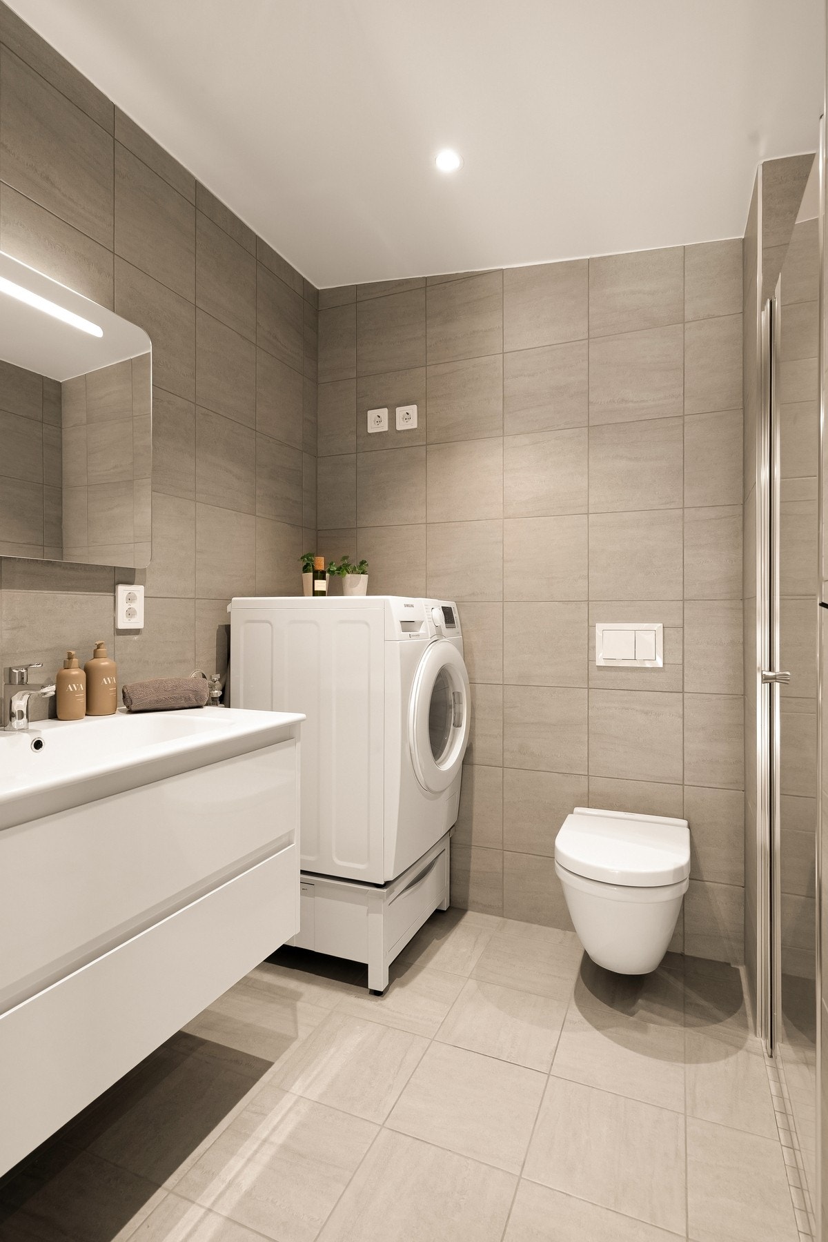 Flislagt bad med varmekabler, dusjnisje, vegghengttoalett, opplegg for vaskemaskin og innredning med vask Galleribilde