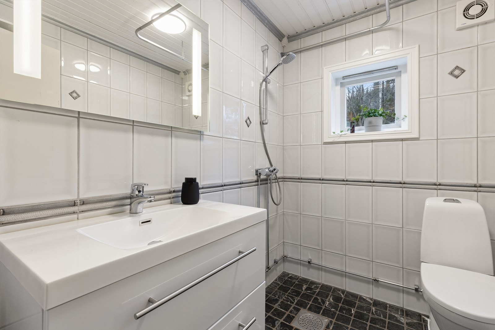 Badet i 1. etasje er fra 2003, iht. tidligere salgsoppgave og har fliser og malte plater på veggene, samt panel i taket. Gulvet er flislagt og utstyrt med elektriske varmekabler. Galleribilde