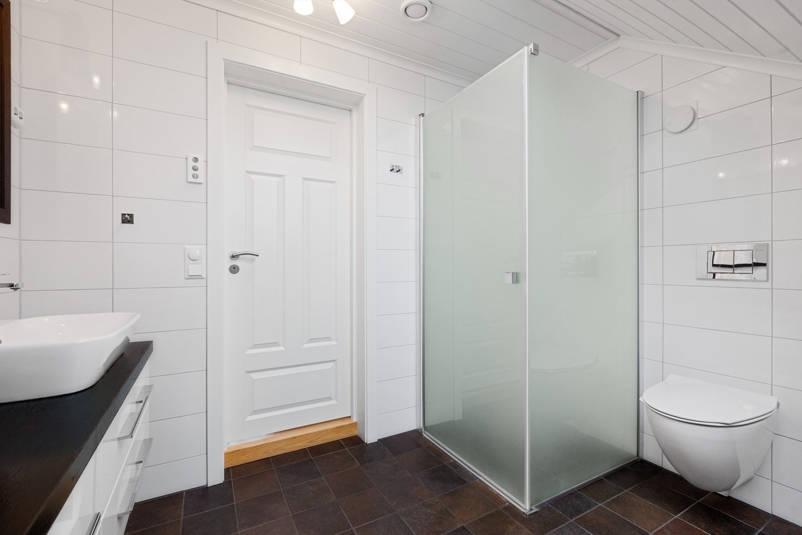 Badet er innredet med en servantinnredning med skuffer, servantskål og speil, et vegghengt toalett og en dusjnisje med glassvegg og dør. Galleribilde