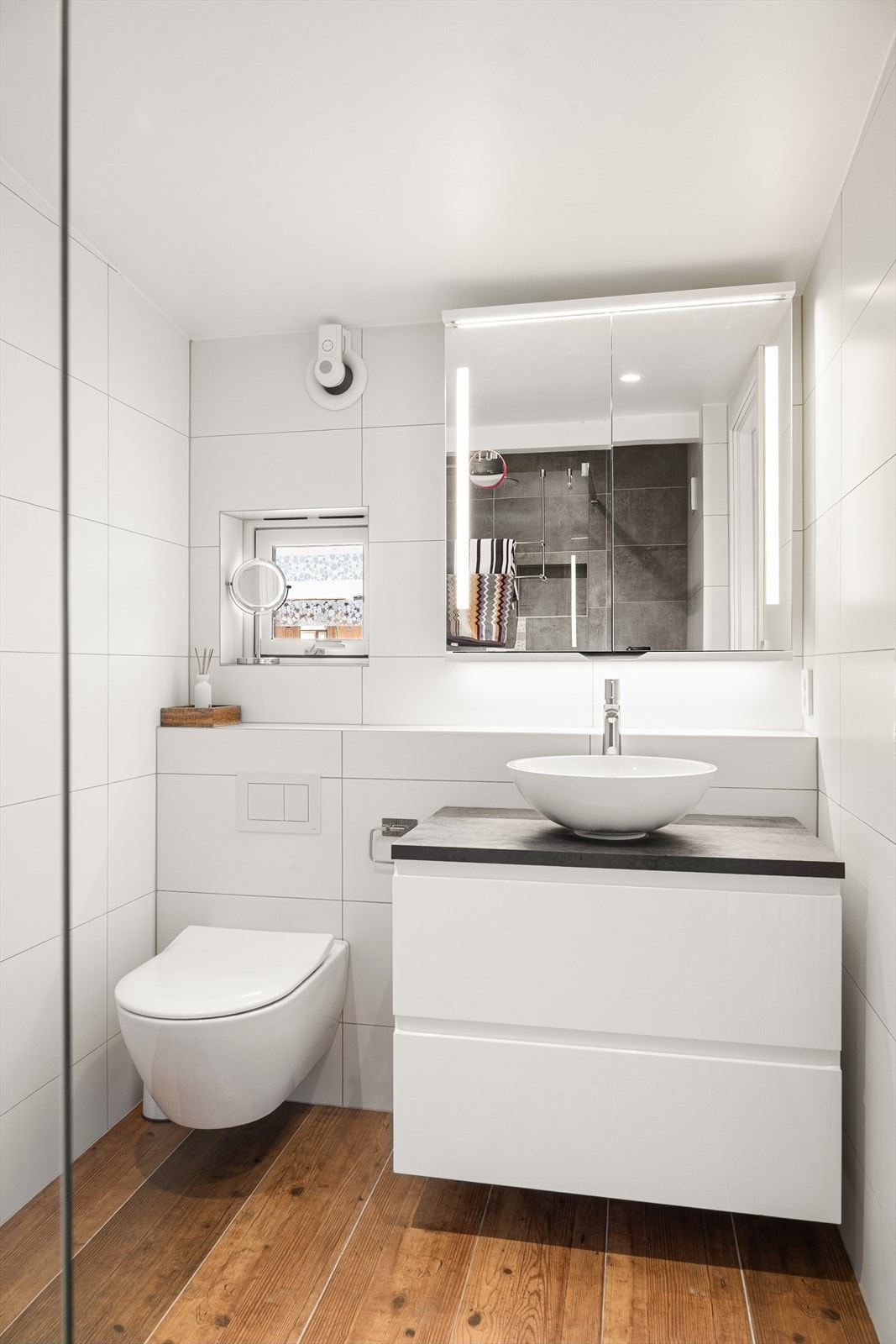 Badet har varme i gulvet. Rommet er innredet med vegghengt servant med underskap, vegghengt toalett og et dusjhjørne med glassvegg. Galleribilde
