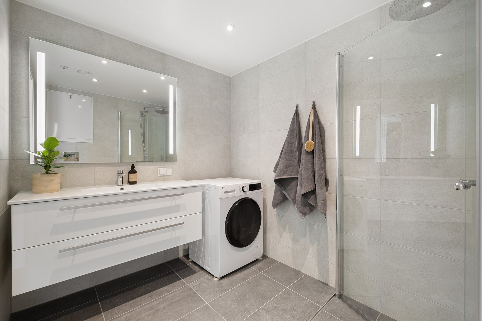 Baderommet inneholder innfellbare og plassbesparende dusjdører i glass, dusjbatteri og garnityr montert på vegg, veggmontert WC og kran og avløp for vaskemaskin. Rørskapet er lokalisert over toalettet. Galleribilde