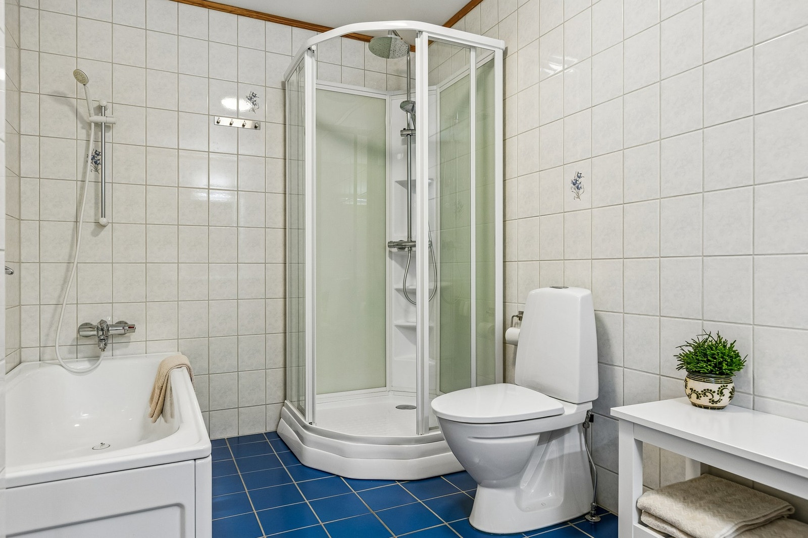 Baderommet er innredet med lyse tidløse fliser på vegger og blå fliser på gulv. Badet er praktisk utstyrt med badekar, dusjnisje og en behagelig varme i gulv. I tillegg til bad, er det i nærhet av entréen et separat toalettrom. Galleribilde