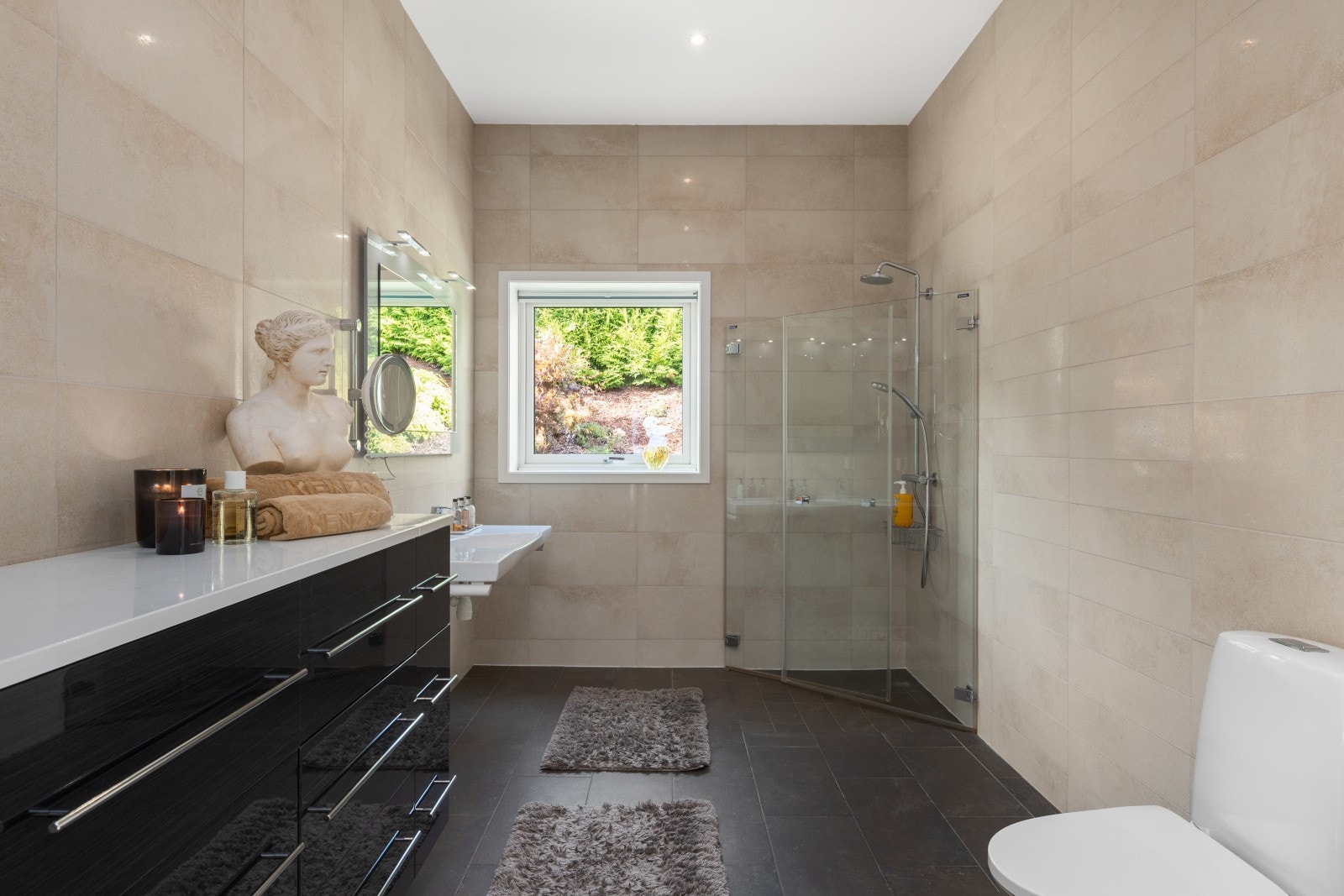 Moderne baderomsinnredning med skuffer og ovenpåliggende benkeplate. Baderommet er også utstyrt med vegghengt servant med speil og belysning over, dusjhjørne med glassdører og både hånd- og regnfallsdusj, samt gulvstående toalett. Galleribilde