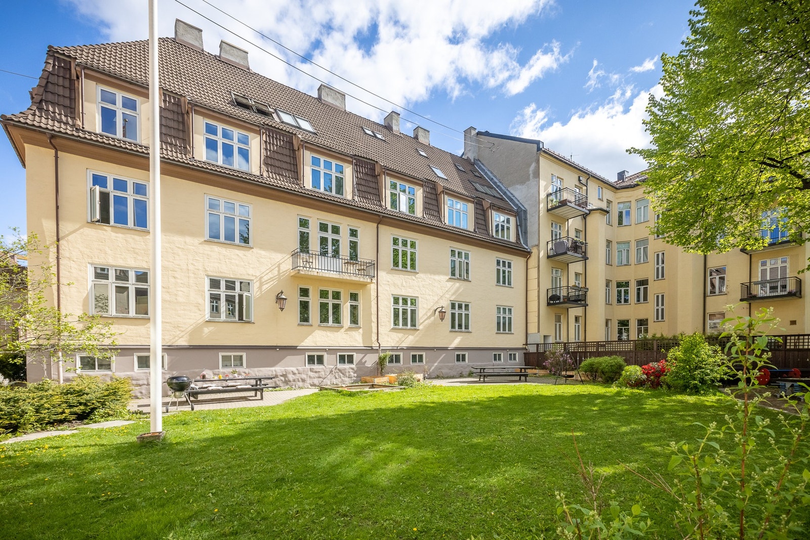 Bygningen er en fire-etasjers leiegård med pusset fasade og skråtak, omgitt av velholdt, grønt fellesområde. Galleribilde