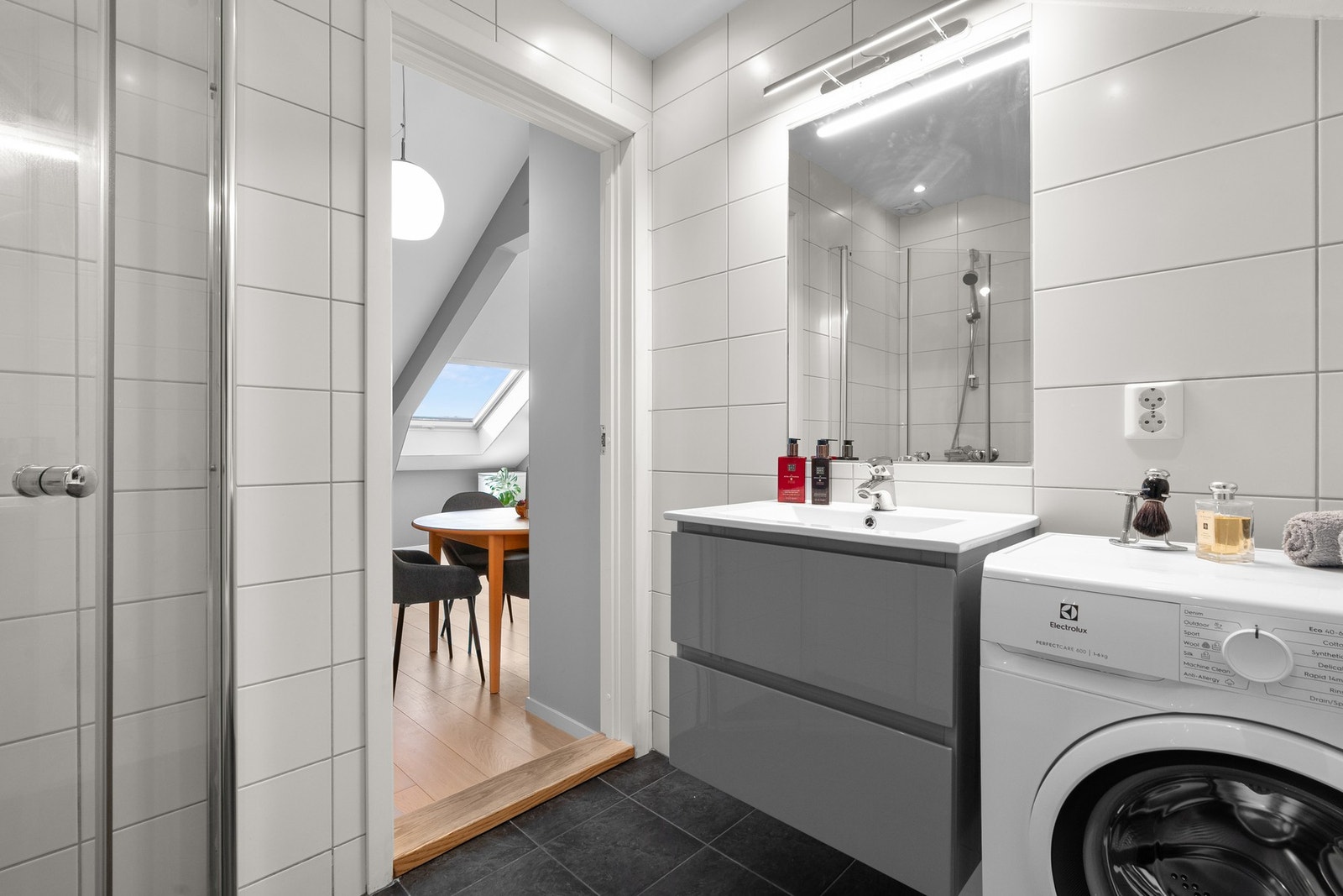 Moderne baderomsinnredning med skuffer og heldekkende servant, speil med belysning over. I hjørnet er det plass og opplegg for vaskemaskin. Galleribilde