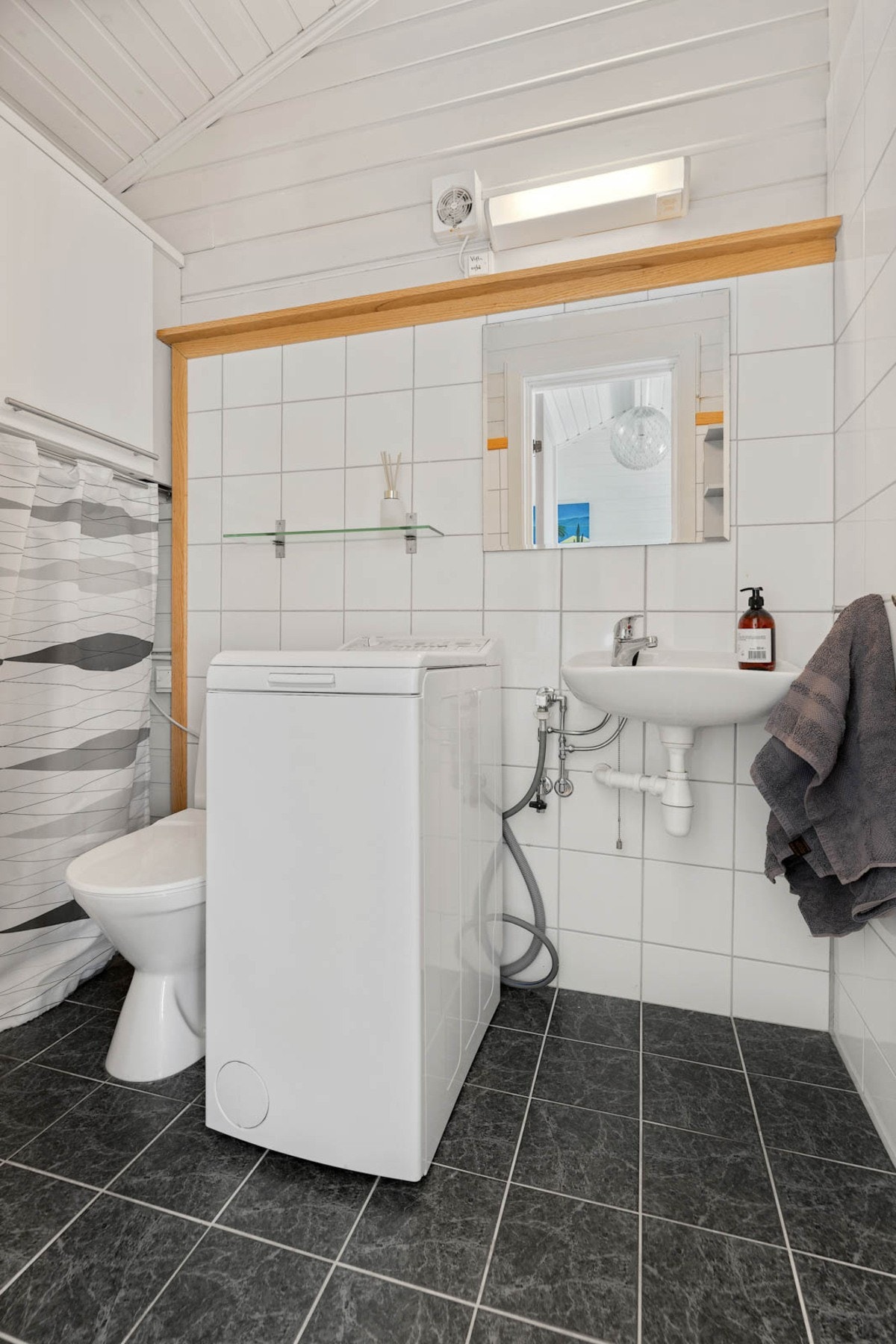 Badet har flislagte vegger og gulv, servant, toalett og vaskemaskin. Galleribilde