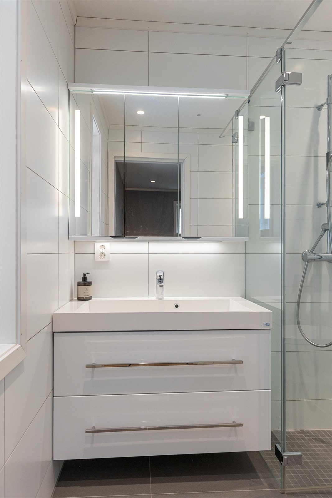 Baderomsinnredning med nedfelt servant, speilskap med lyslist, veggmontert toalett med spylefunksjon samt dusj med glassvegger. Galleribilde