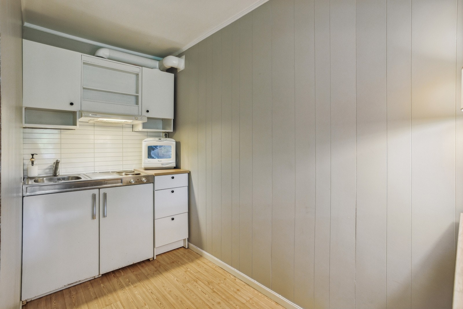 Enkelt hybelkjøkken med lyse slette fronter og laminert plate på vegg over vask og benk. Galleribilde