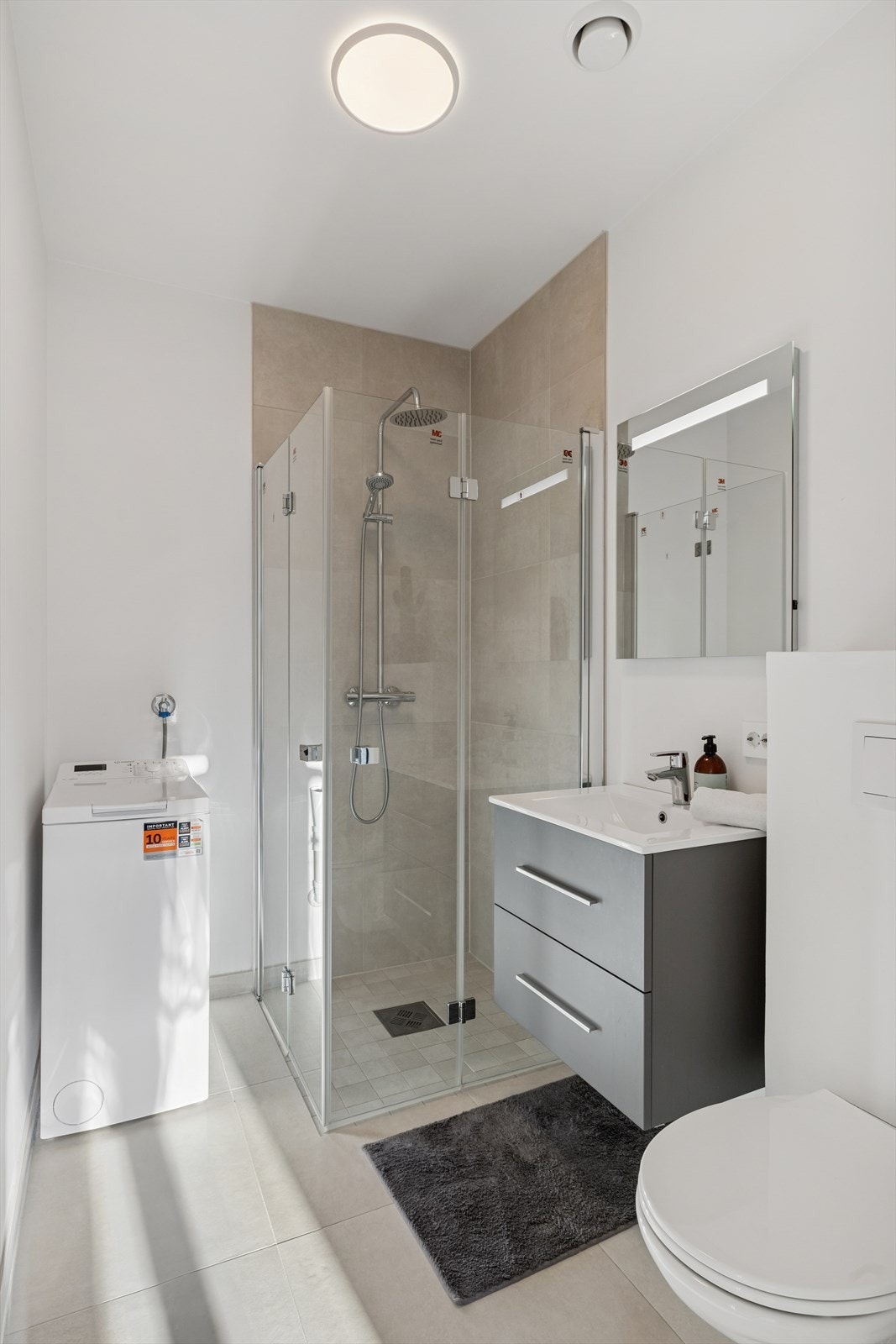 Tidløst kombinert bad og vaskerom med flislagt gulv, baderomsinnredning, vegghengt toalett, dusjhjørne og opplegg for vaskemaskin. Galleribilde