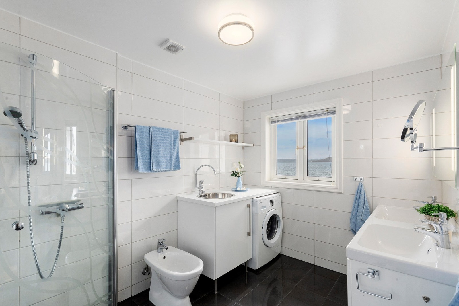 Badet fremstår som lyst og moderne med flislagte overflater, dobbelt servant, romslig dusjhjørne, praktisk opplegg for vaskemaskin, toalett og bidet. Galleribilde