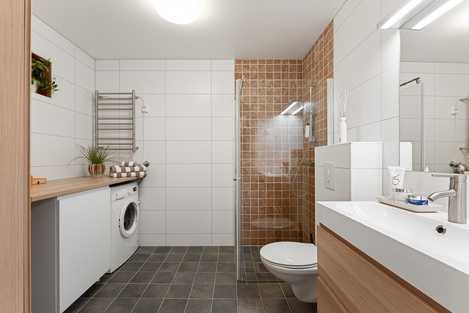 Pent baderom med fliser på gulv med gulvvarme, fliser på vegger og malt himling. Badet har vegghengt toalett, vegghengt servantinnredning, speil med overlys og dusjhjørne med dører Galleribilde