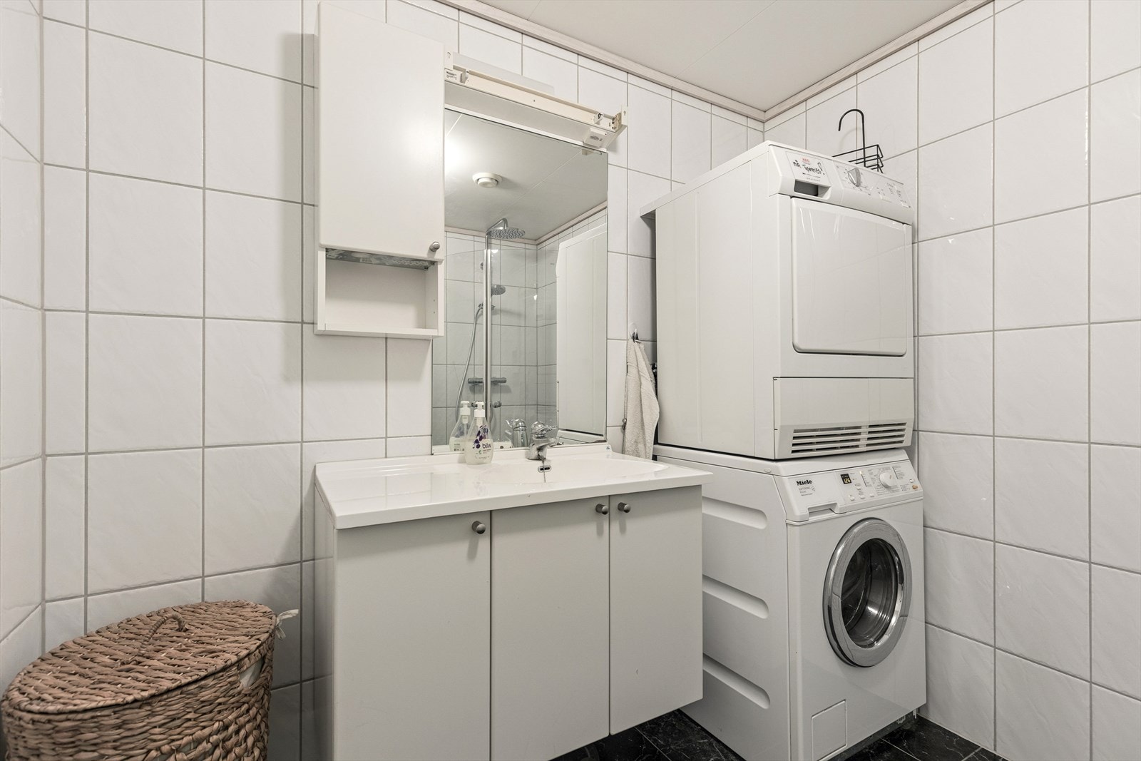 Pent fliselagt baderom med servant med møbel, toalett, opplegg for vaskemaskin og dusj. Galleribilde