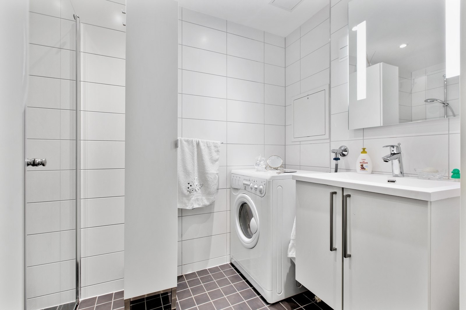 Badet er helfliset og utstyrt med dusjhjørne med glassvegger, vegghengt toalett og opplegg for vaskemaskin. Baderomsinnredning med servant og god oppbevaringsplass i skap. Galleribilde