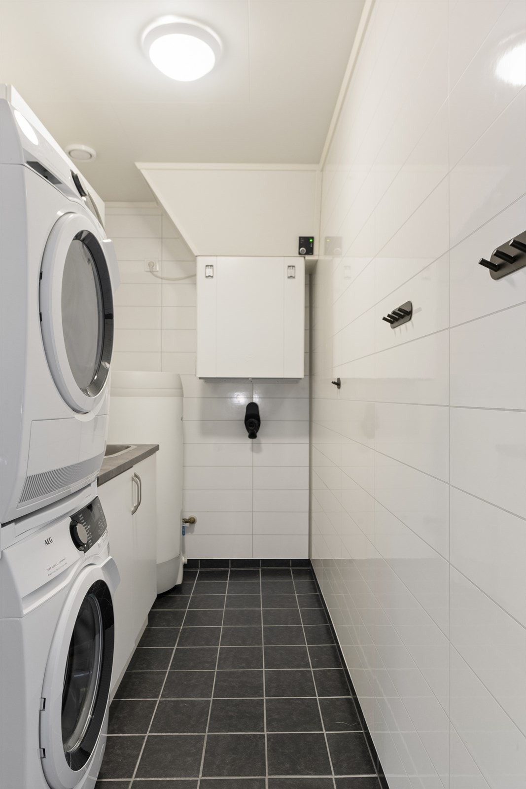 Vaskerom hvor man har fliser på gulv og baderomsplater på vegger. Her har man skap til oppbevaring, opplegg for vaskemaskin og utslagsvask. Galleribilde