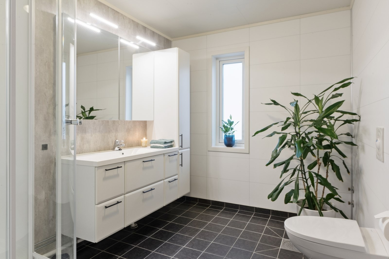 Bad med fliser på gulv og baderomsplater på vegger. Her har man vegghengt toalett, høyskap, dusjkabinett, baderomsinnredning med servant og overhengende speil med belysning og lagringsplass. Galleribilde