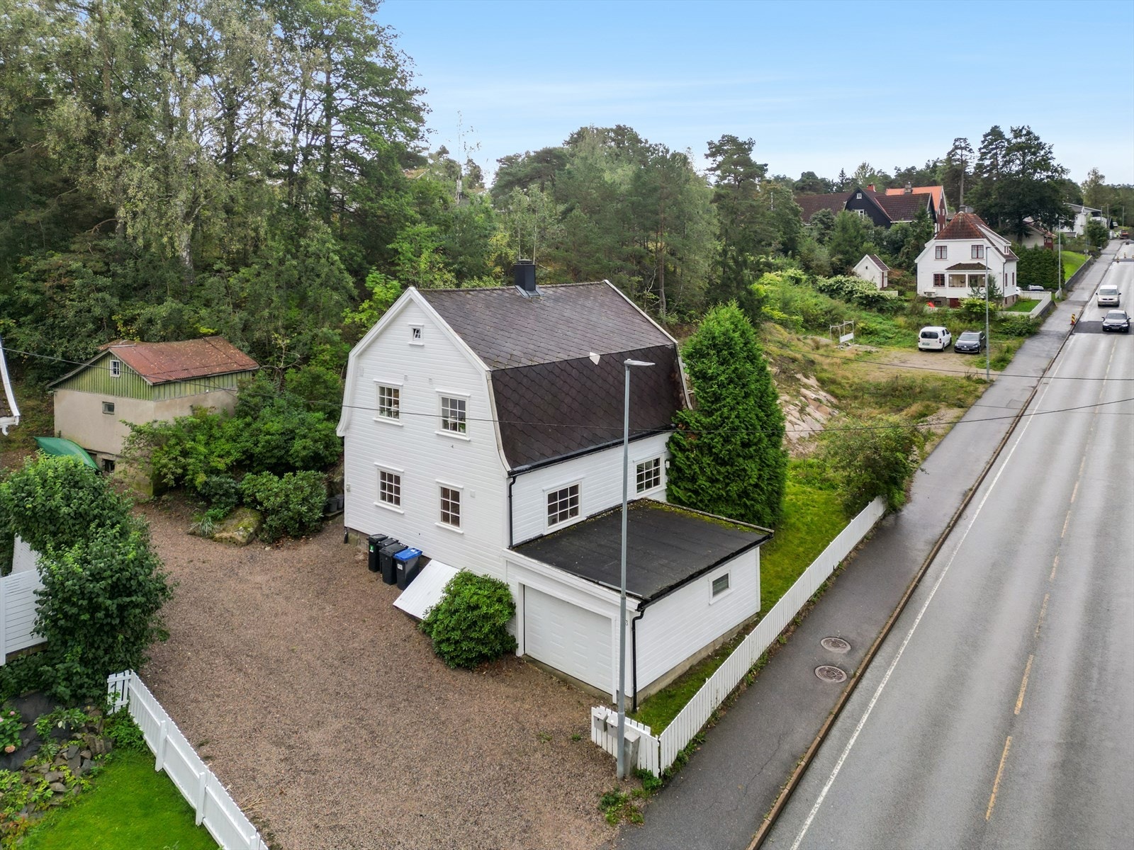 Gruset innjørsel og gårdsplass, garasje er bygget inntil boligen. Tomtegrensen går et stykke opp i terreng bak stallbygning til venstrte i bildet. Galleribilde