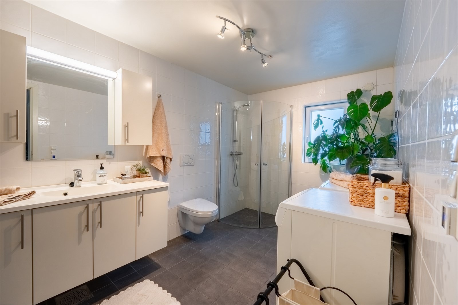 Innbydende bad med hvit baderomsinnredning, vegghengt toalett, dusjhjørne og opplegg for vaskemaskin Galleribilde