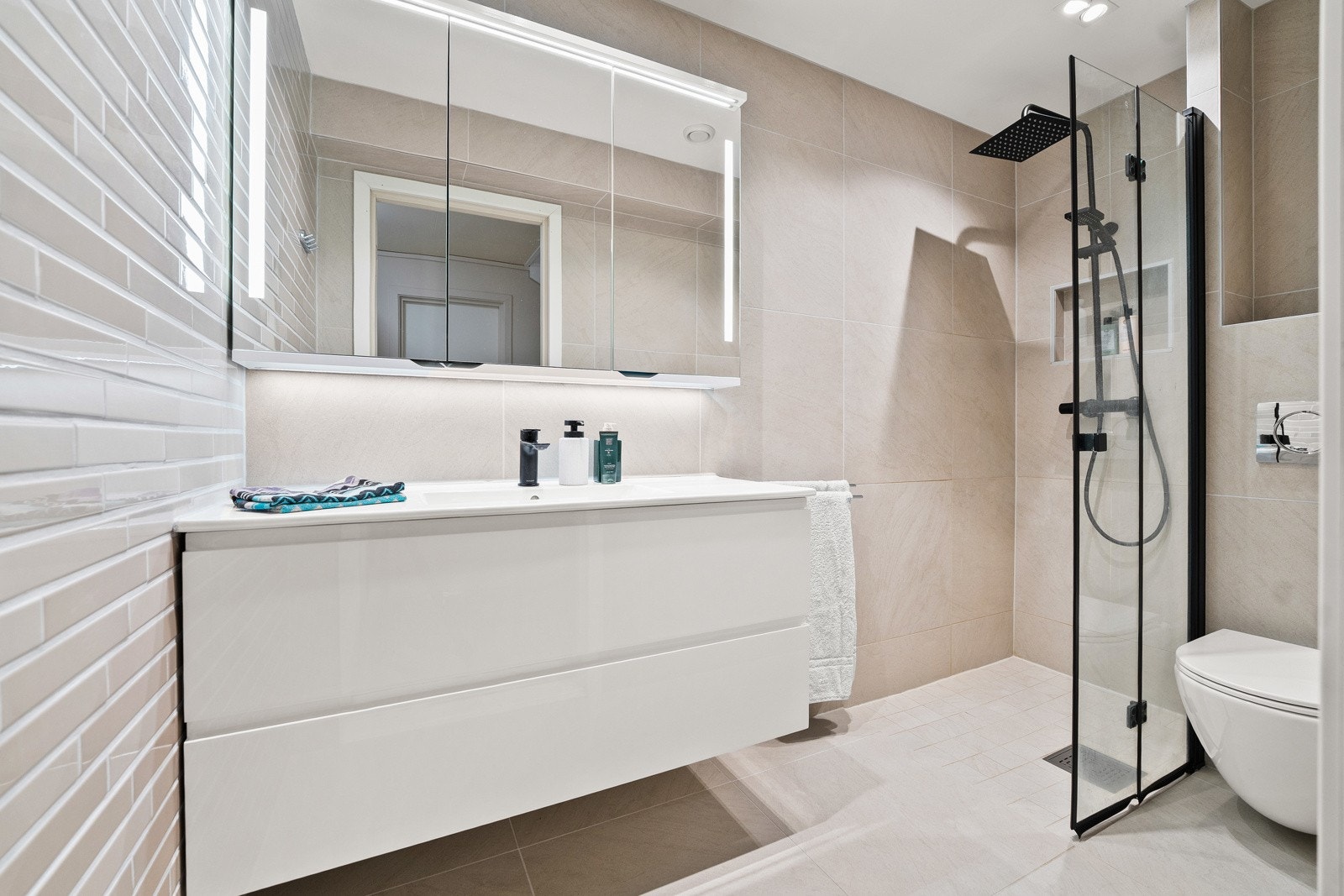 Badet fremstår som lyst og praktisk med flislagte overflater på vegger og gulv. Dusj, toalett og servant med underskap i hvite fronter. Galleribilde