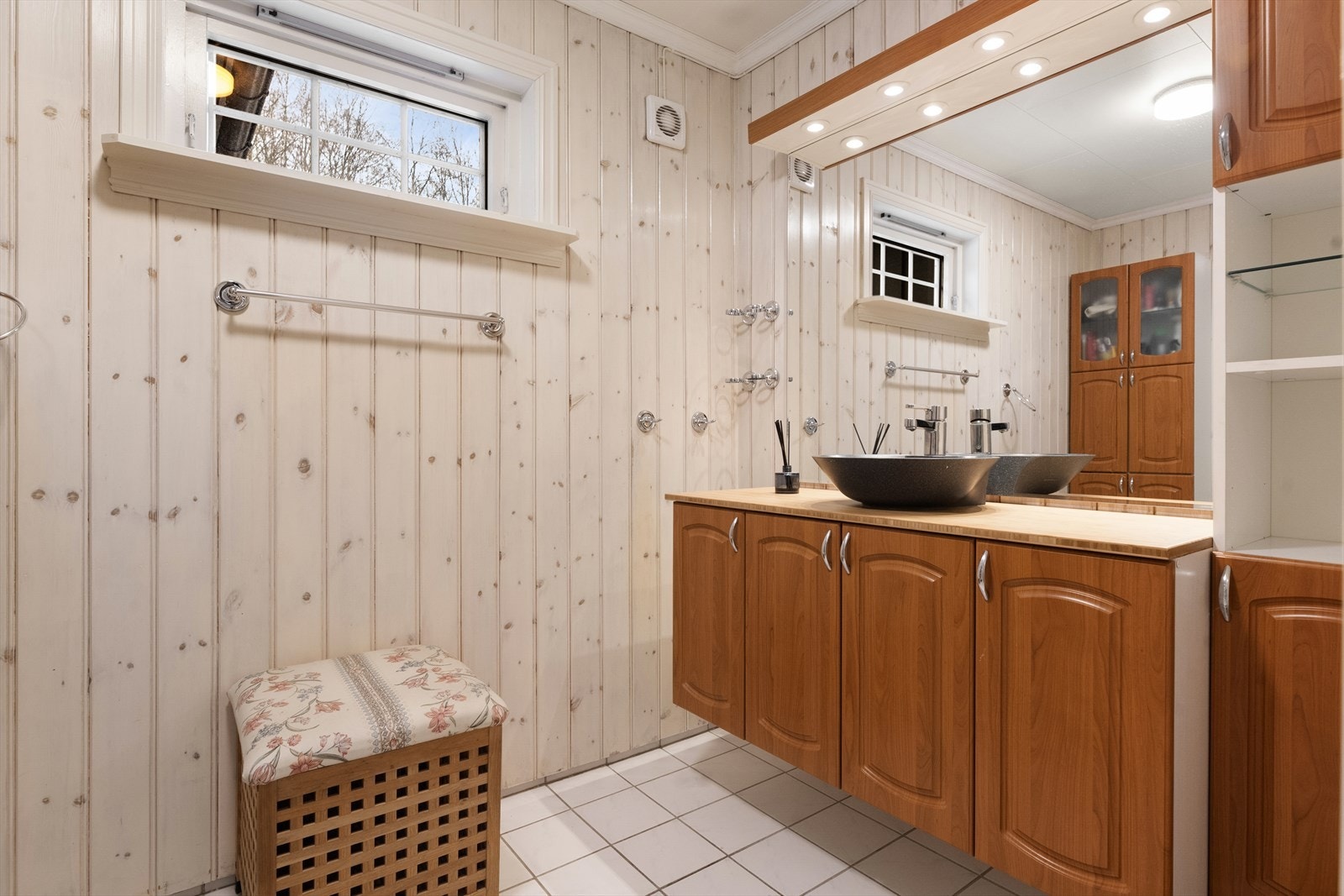 Badet er innredet med dusjhjørne med glassvegger og servantinnredning med skap, hyller og speil med overlys. Det er elektrisk avtrekksvifte på vegg. Galleribilde