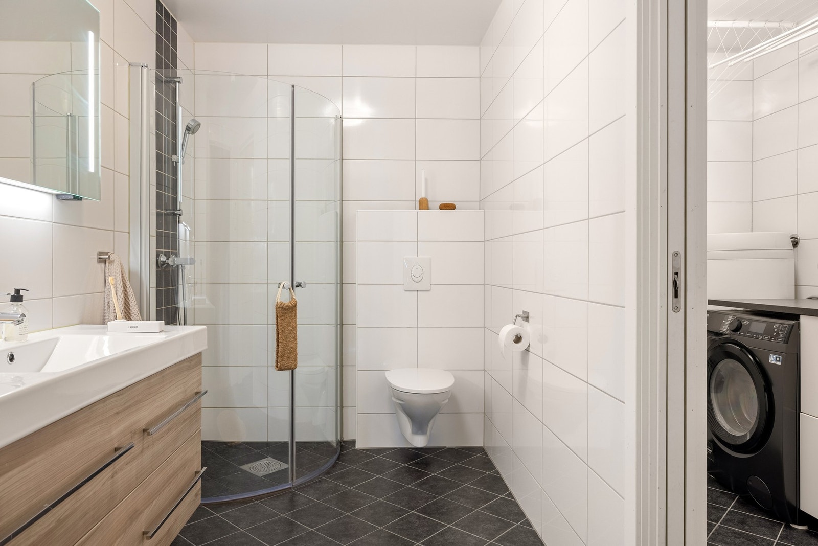 Badet er innredet med vegghengt toalett, dusj med glassvegger, og baderomsmøblement med speil og belysning. Galleribilde