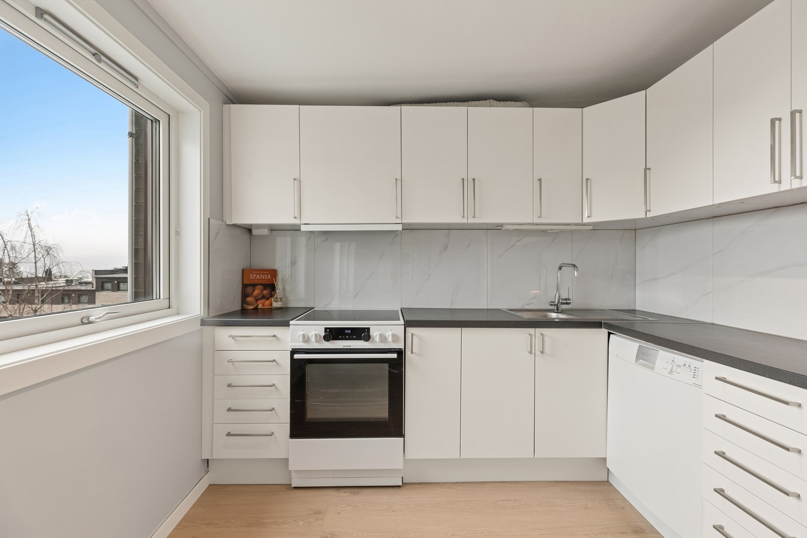 Kjøkken ligger i en delvis åpen løsning mot stuen og er innredet med hvite, glatte fronter og en mørkere laminat benkeplate som gir et kjøkkenet et moderne og tidsriktig preg. Galleribilde