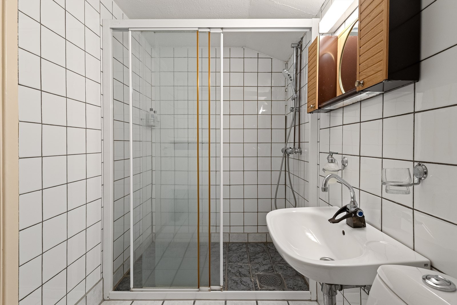 Badet i kjelleren inneholder dusj, toalett og servant med skap og speil over. Innredningen avviker fra byggesøkte tegninger - rommet er ikke byggesøkt/godkjent. Galleribilde