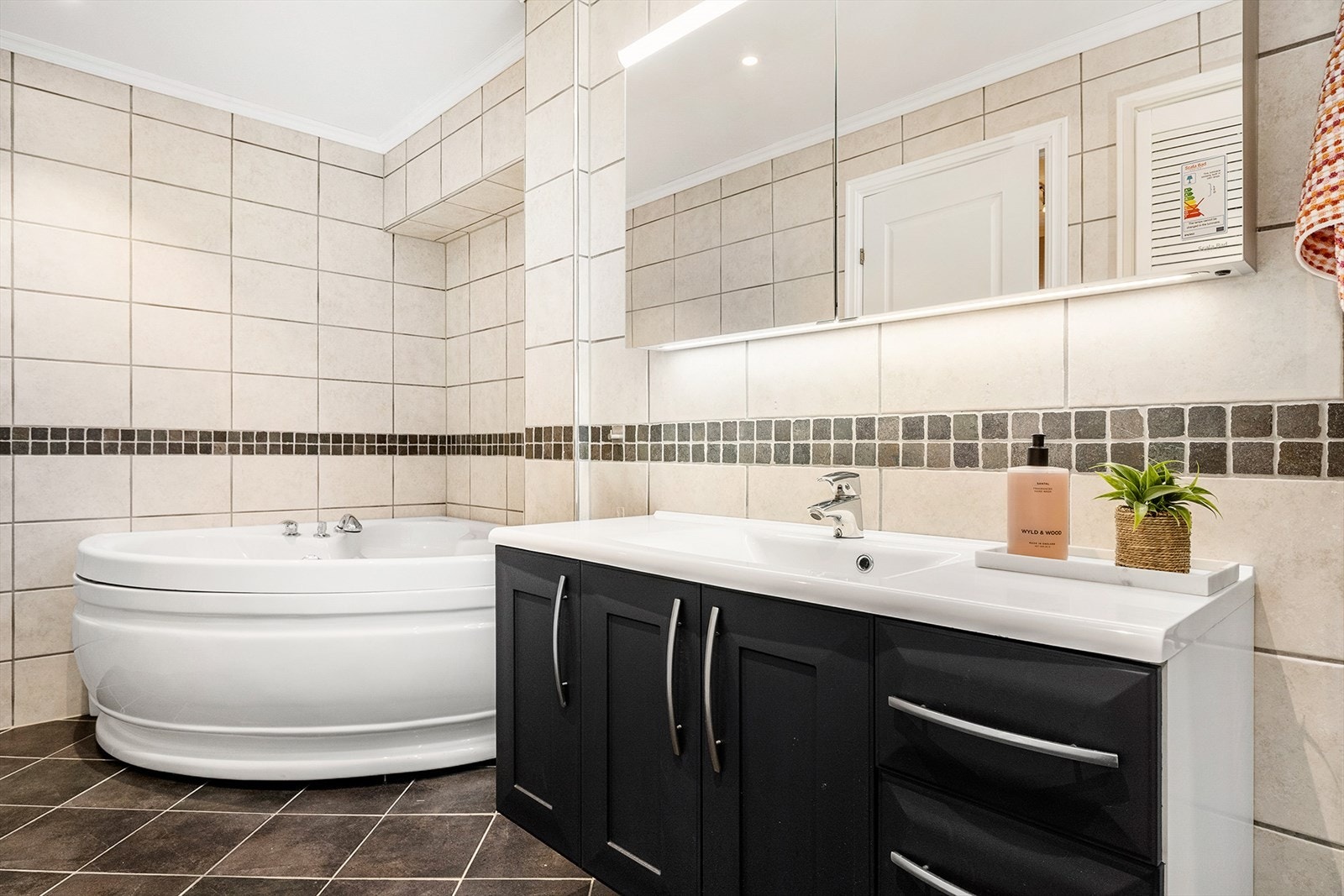 Badet er innredet med veggfestet WC, dusjkabinett, hjørnebadekar og baderomsinnredning med speilskap. Skjult bak dører er det vaskeromsavdeling under trappen. Galleribilde