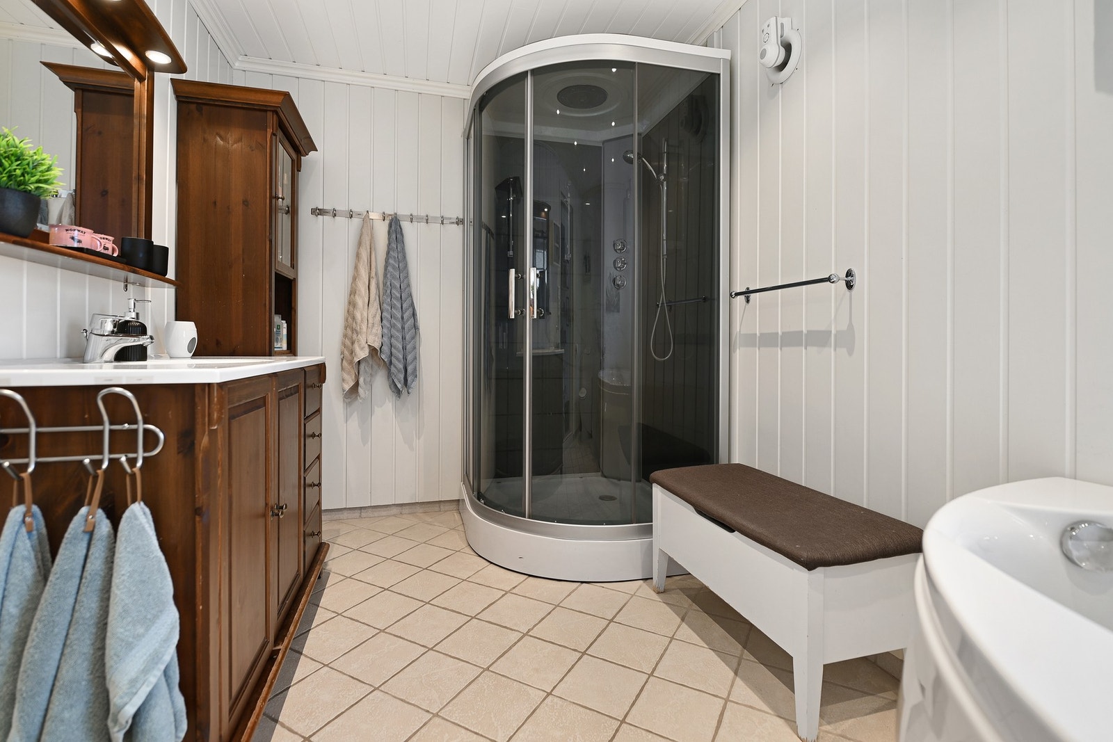 Badet har dusjkabinett, servant med speil, og god oppbevaringsplass. Veggene er hvite og gulvet har lyse fliser. Galleribilde