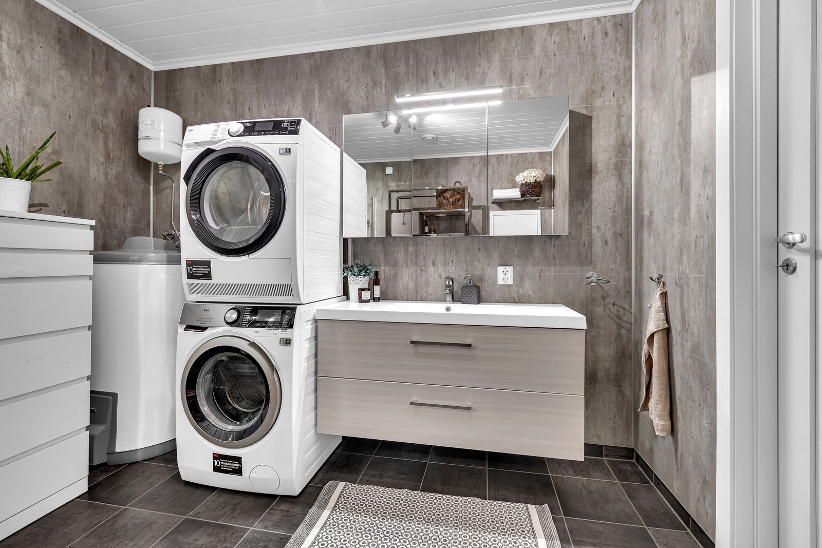 Baderomsinnredningen med skuffer og speilskap med lys. Videre har baderommet gulvmontert toalett, dusjhjørne med innfellbare glassdører og opplegg for vaskemaskin og tørketrommel. Galleribilde